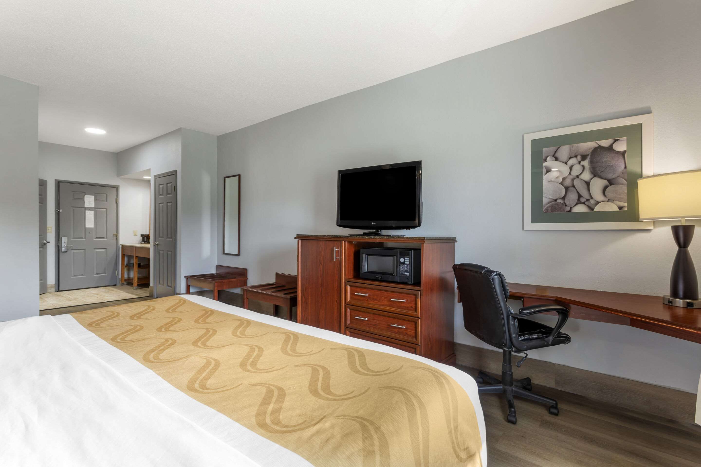 A hotel room with a bed, a desk with a chair, a television, a microwave, and a framed picture on the wall.
