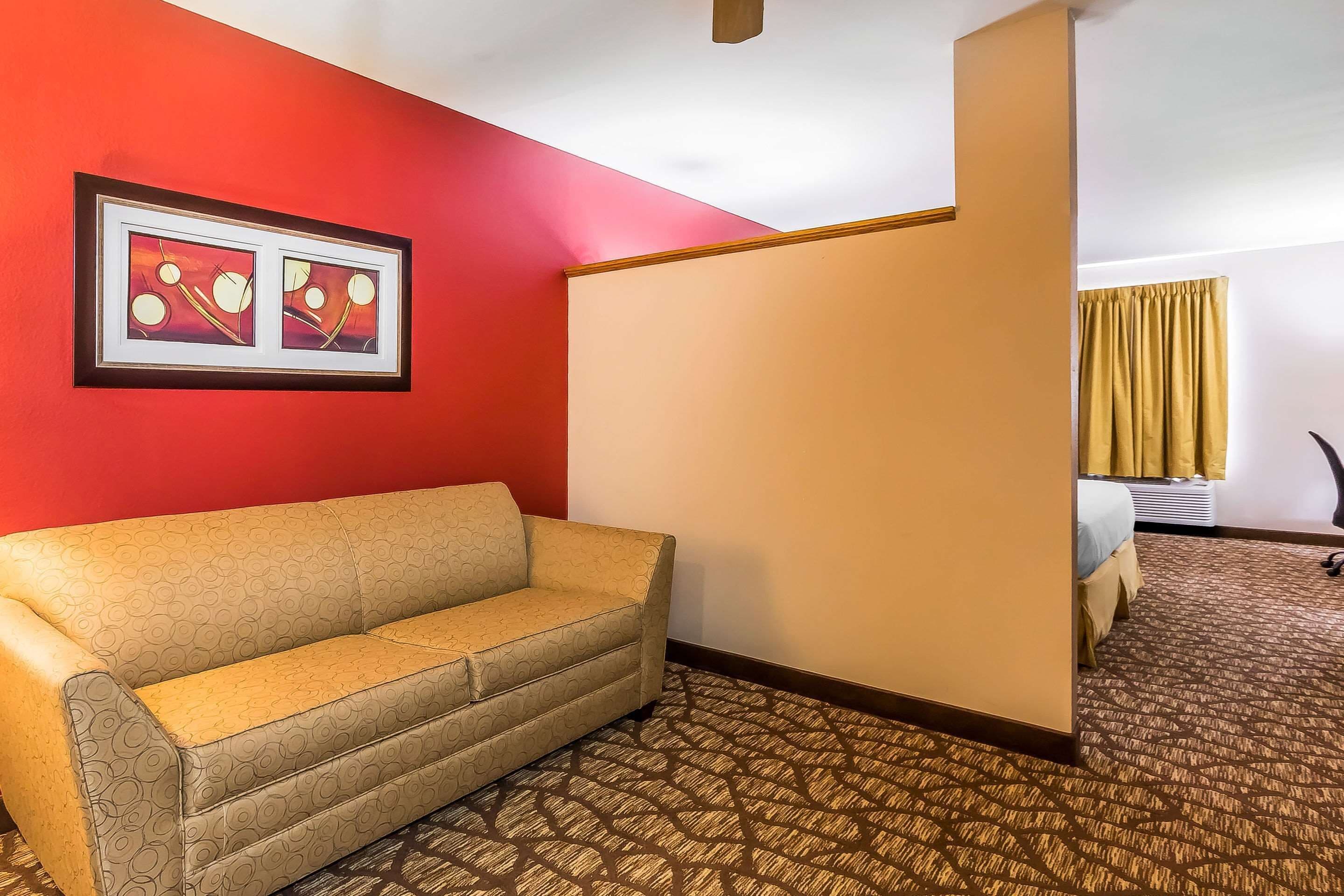 A hotel room with a beige sofa, a red accent wall, a framed artwork, a bed with a yellow curtain, and a patterned carpet.