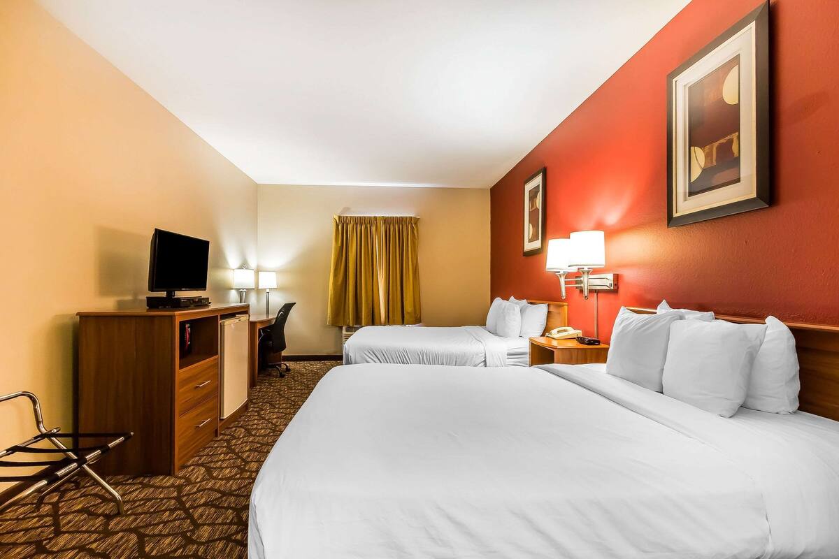 A hotel room with two beds, a desk with a television, a chair, a desk lamp, and a nightstand with a lamp.