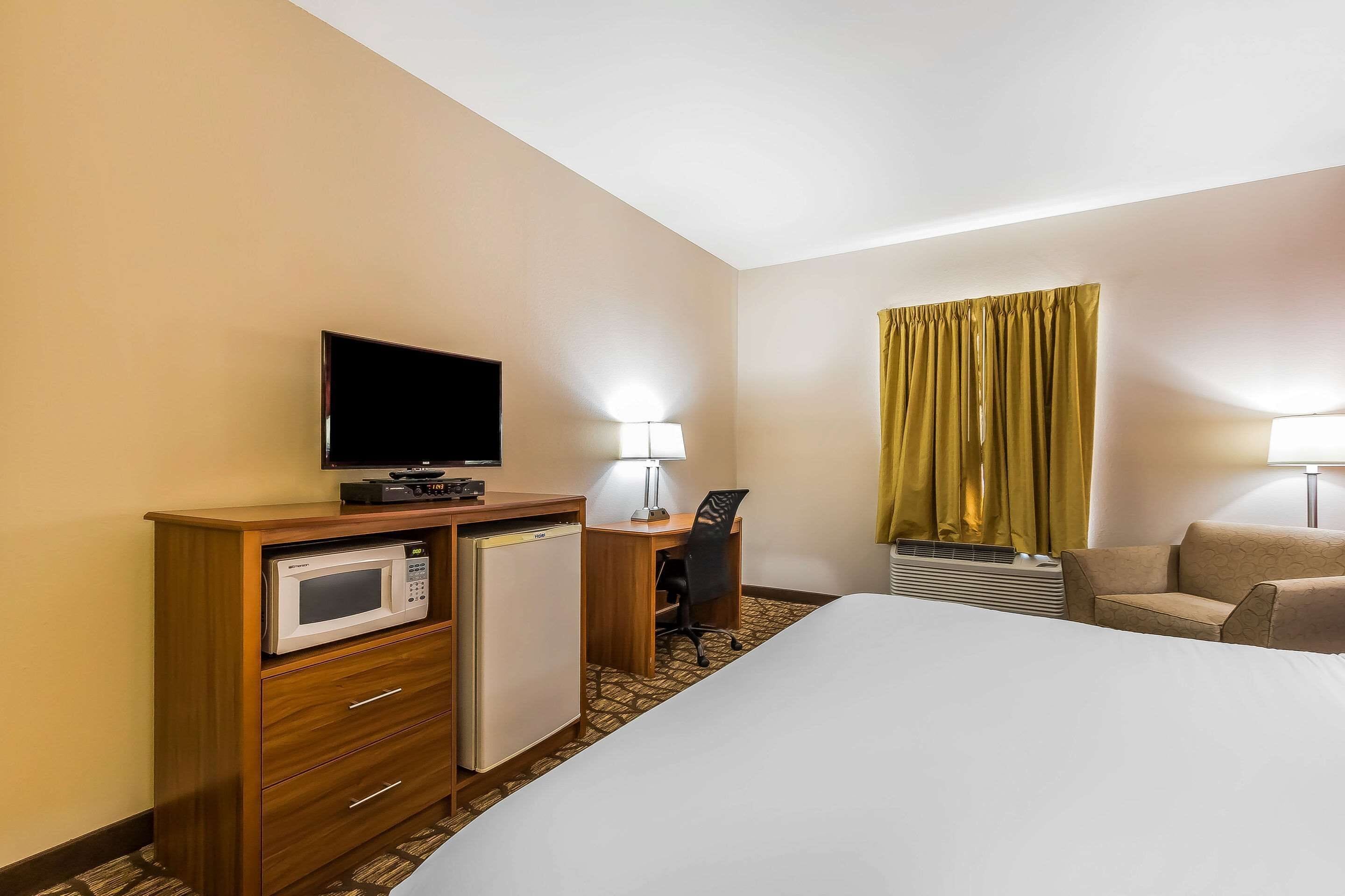 A hotel room with a bed, a desk with a microwave, a TV, a lamp, a chair, and a window with curtains.