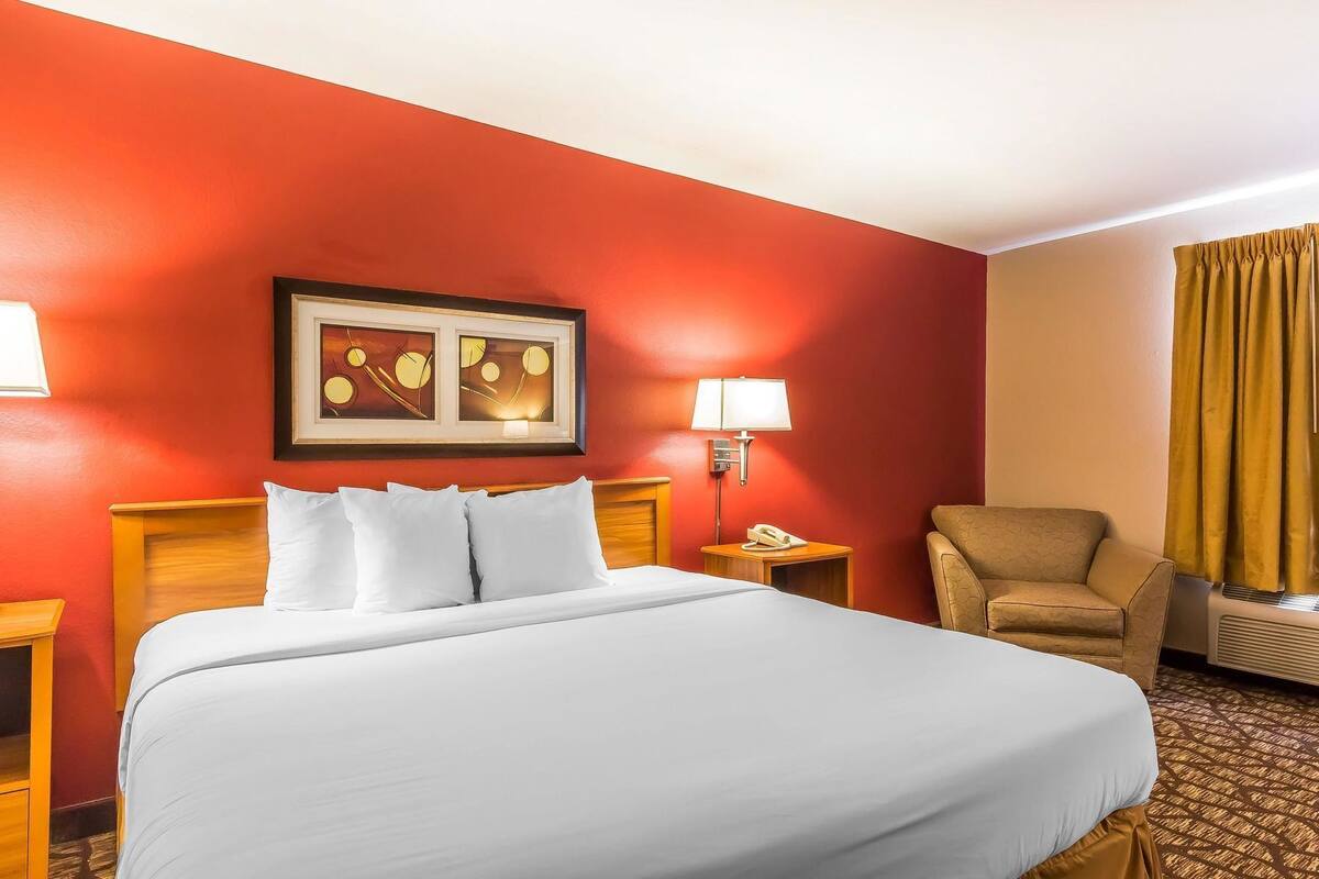 A hotel room with a large bed, a chair, a desk with a lamp, and a framed picture on the wall.
