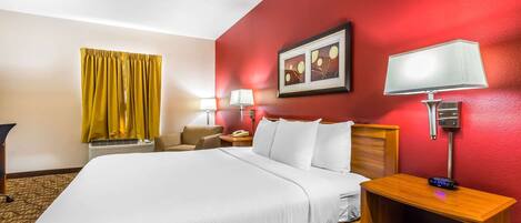 A hotel room with a large bed, a wooden nightstand, a desk, a chair, a lamp, and a picture on the wall.