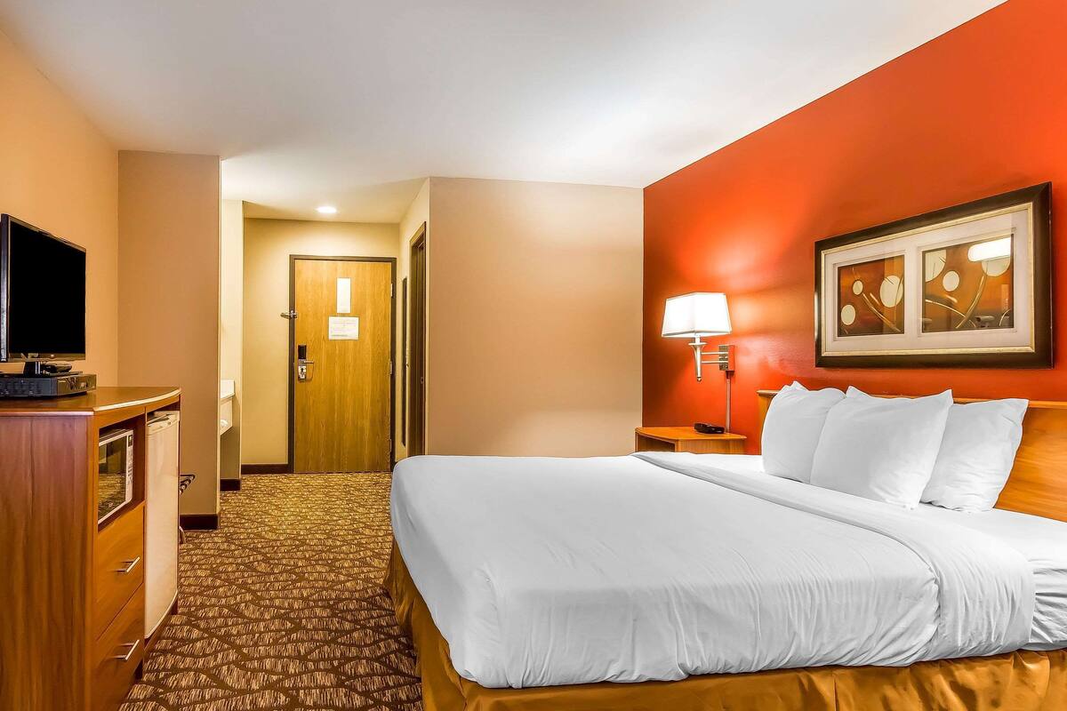 A hotel room with a large bed, a desk with a television, a door, and a wall-mounted lamp.