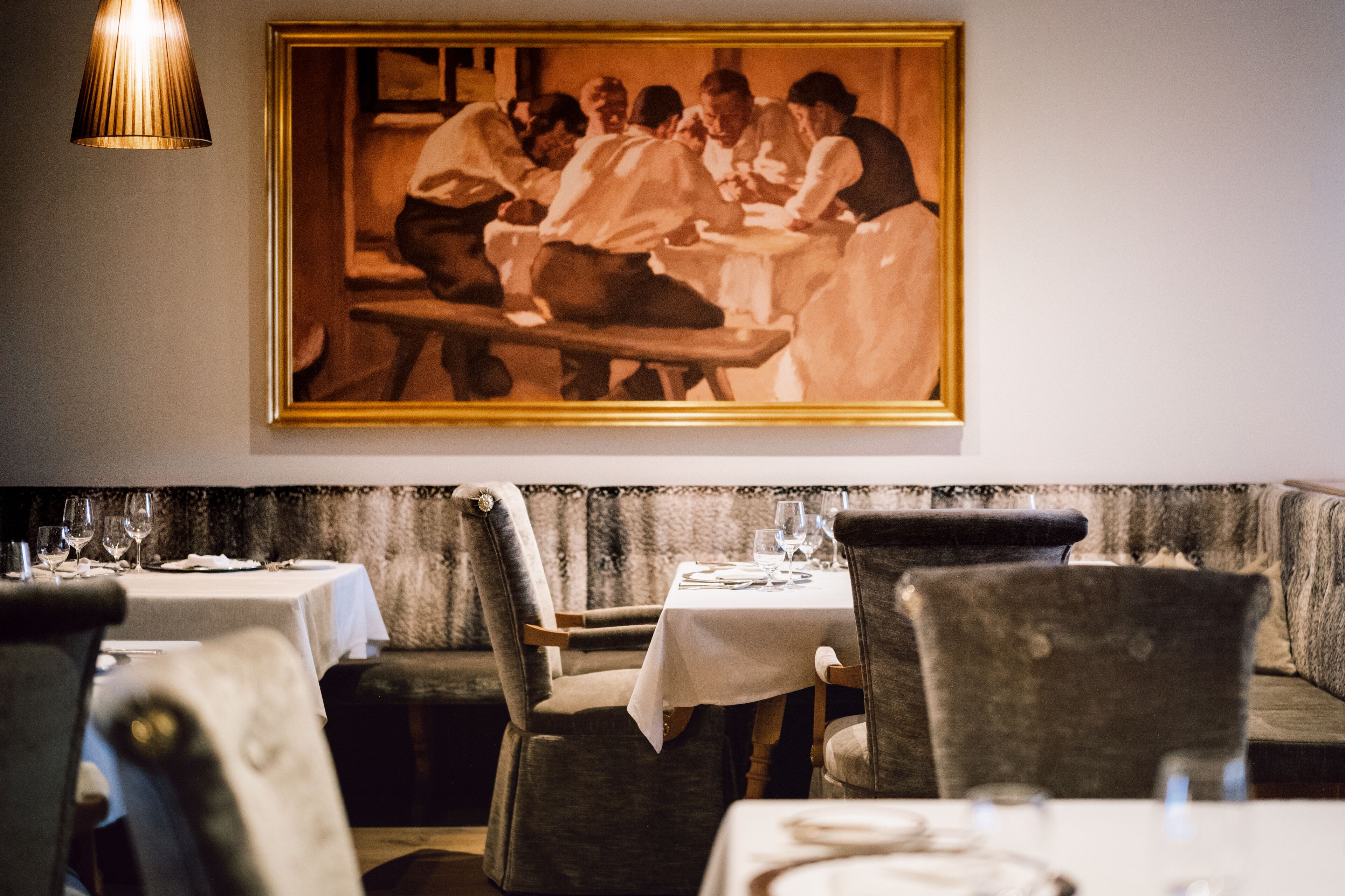 A restaurant interior with a dining table set for two, a framed historical painting on the wall, and a cushioned bench seating area.