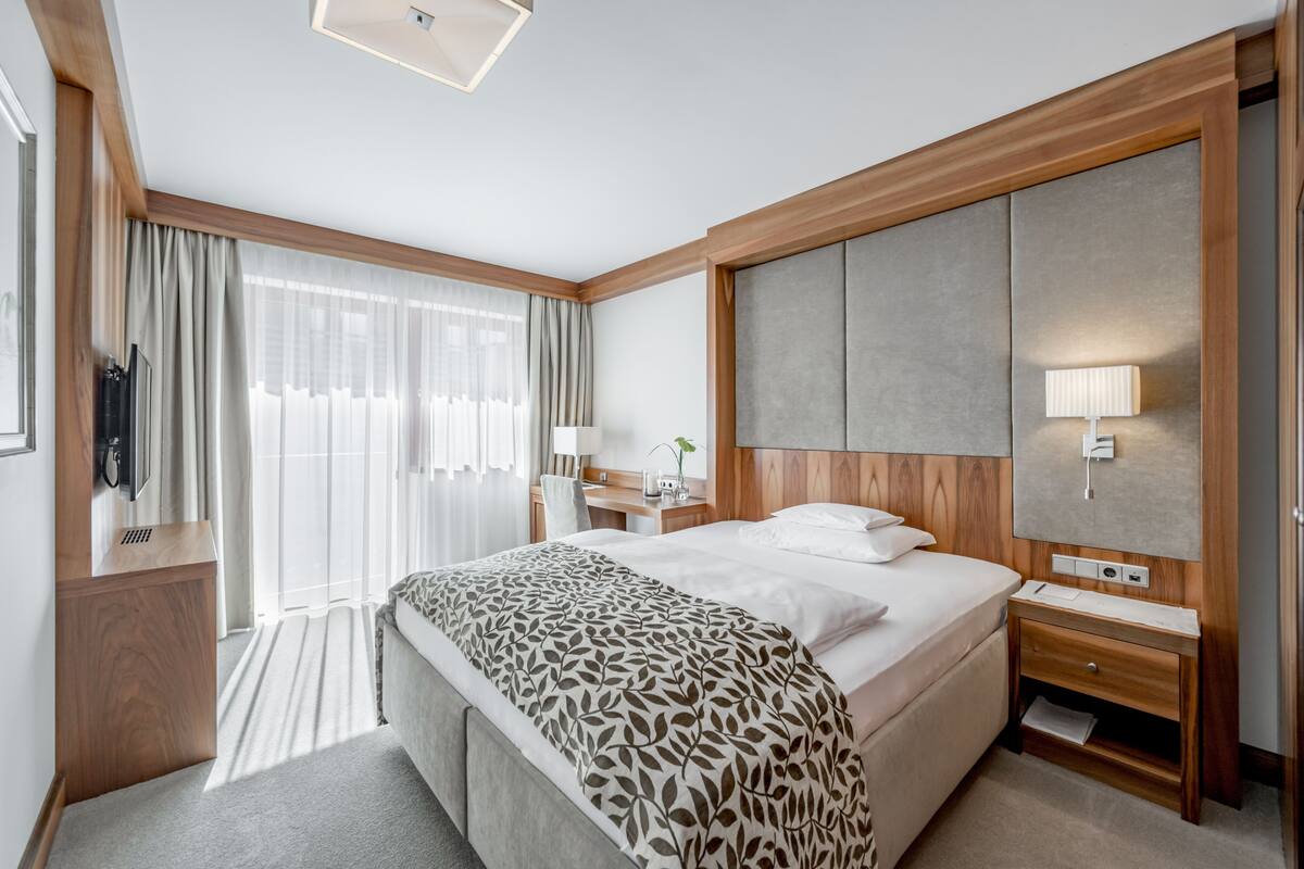 A modern hotel room with a large bed, a wooden headboard, a desk with a lamp, and a TV on a wooden stand.