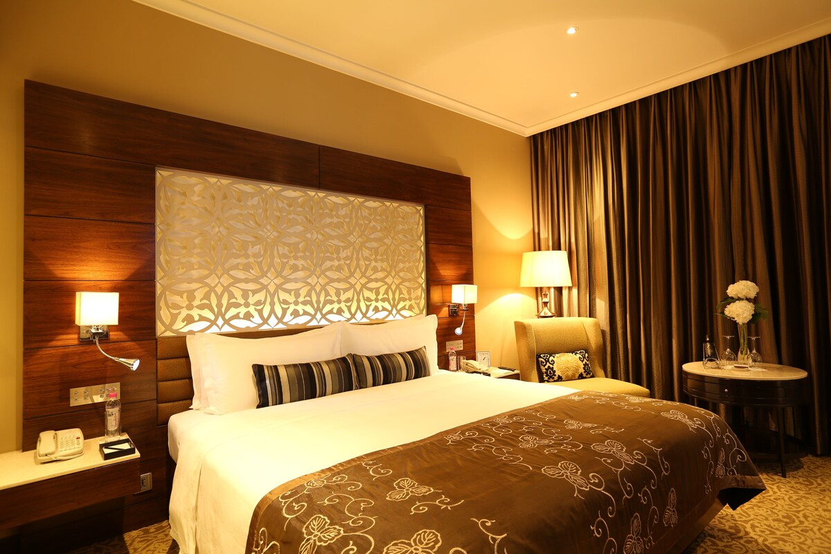 A hotel room with a large bed, a bedside table with a lamp, a telephone, and a decorative headboard.