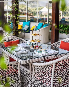 A glass-top table with a tiered tray holding various pastries, surrounded by wicker chairs with red cushions.