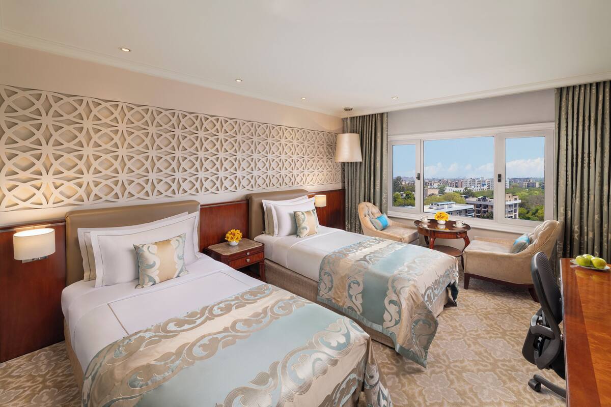 A hotel room with two beds, a desk, a chair, and a large window offering a city view.