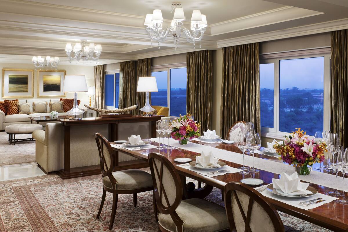 A spacious dining area with a long table set for a meal, a sofa set in the background, and a view of the outdoors through large windows.