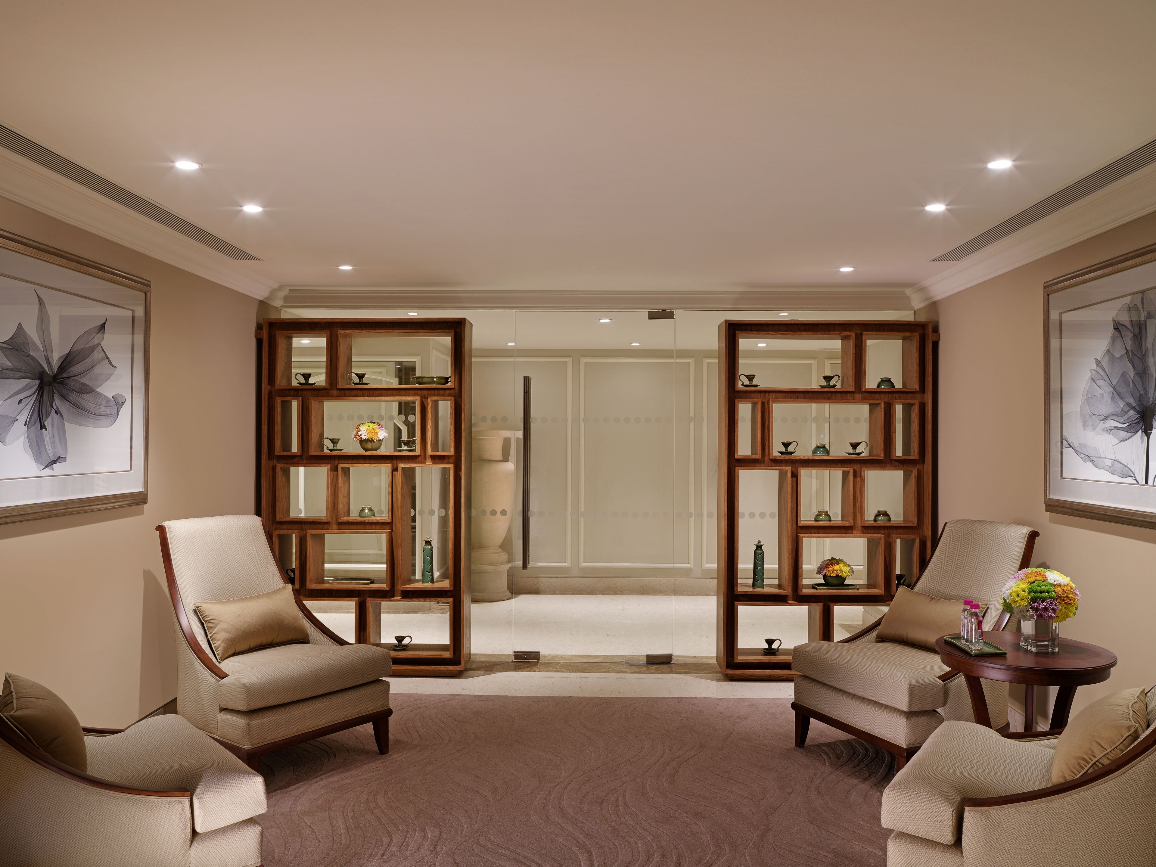 A spacious room with two armchairs, a round table with a vase of flowers, and a wooden cabinet with glass doors displaying various items.