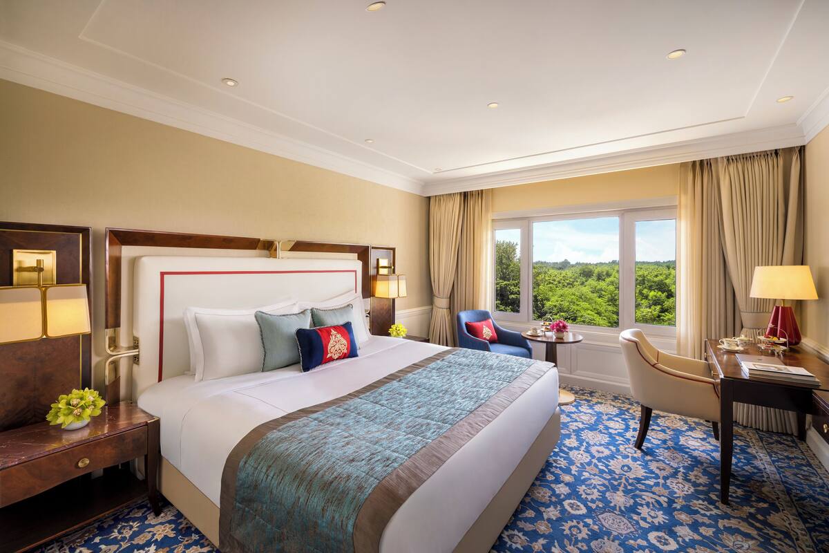 A hotel room with a large bed, a desk, a chair, a window with a view, and a nightstand with a lamp.