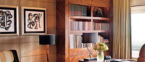 A modern hotel room with a wooden accent wall, a bookshelf, a dining area with a table and chairs, and a large window with curtains.