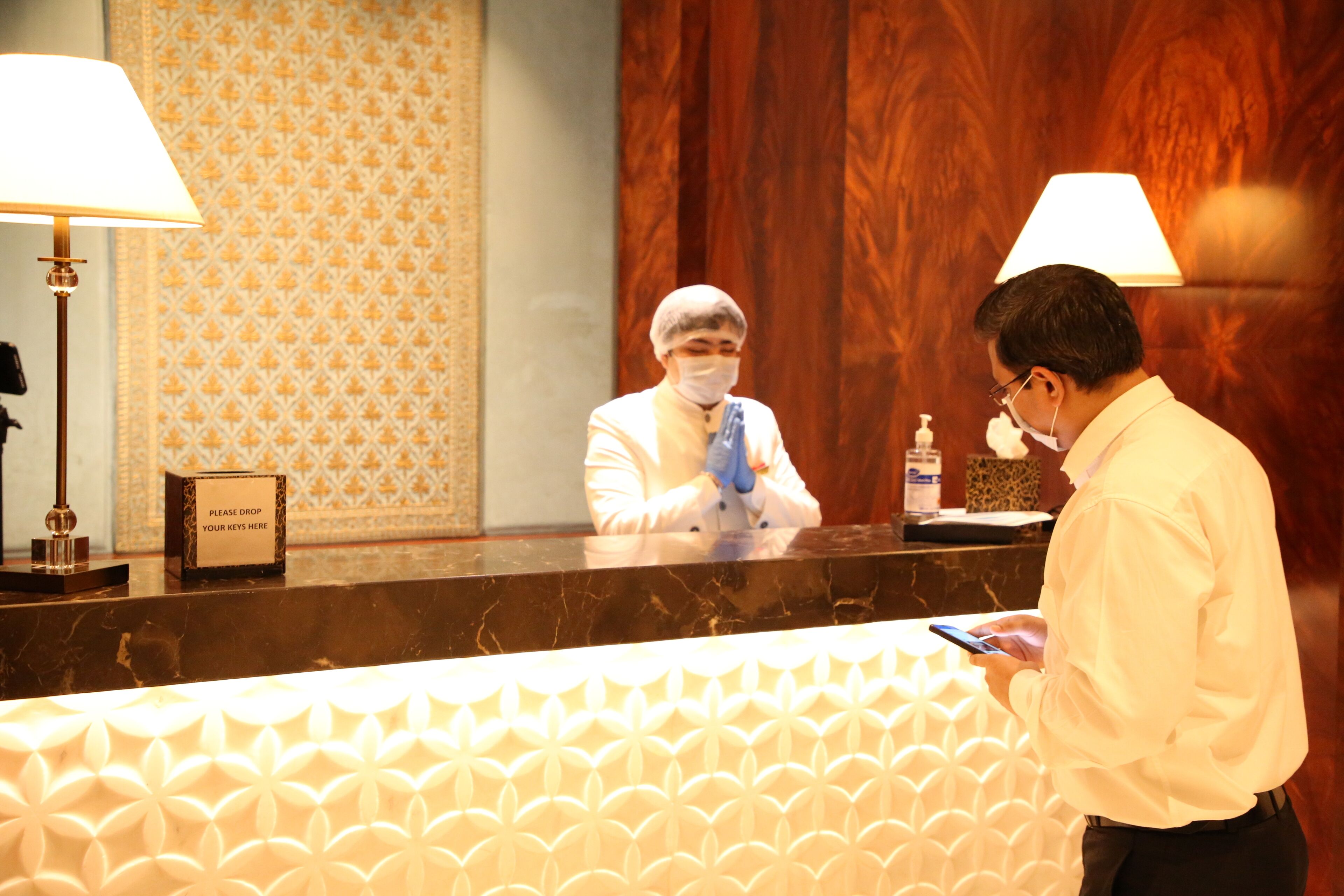 A hotel reception desk with a patterned wall and wooden paneling. A reception desk with a sign reading "Please drop your keys here" is visible.
