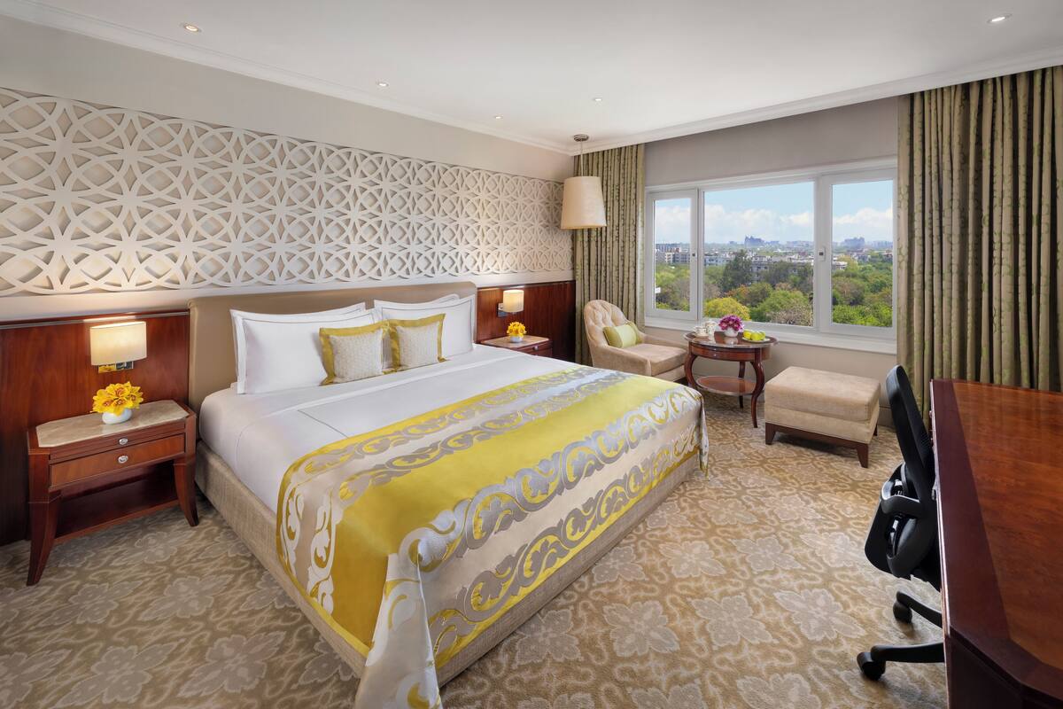 A hotel room with a large bed, a desk, a chair, a nightstand with a lamp, and a view of the city through the window.