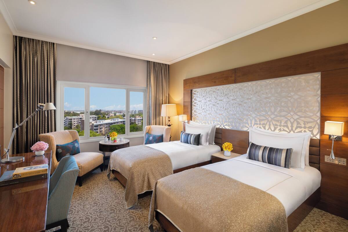 A hotel room with two beds, a desk, and a chair. There is a window with a view of buildings outside.