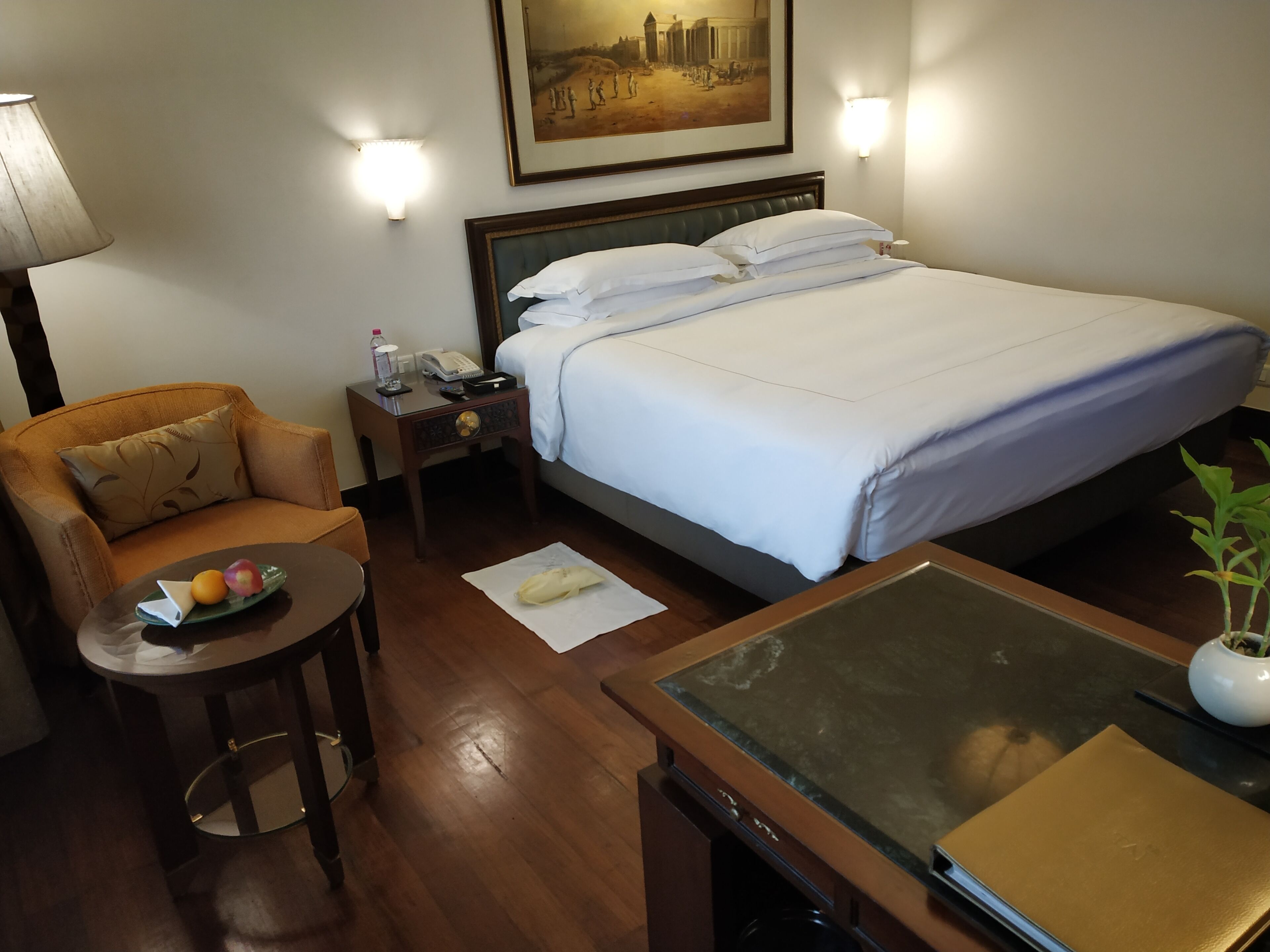 A hotel room with a bed, a chair, a small table with a bowl of fruit, a lamp, and a nightstand with a phone and a bottle.