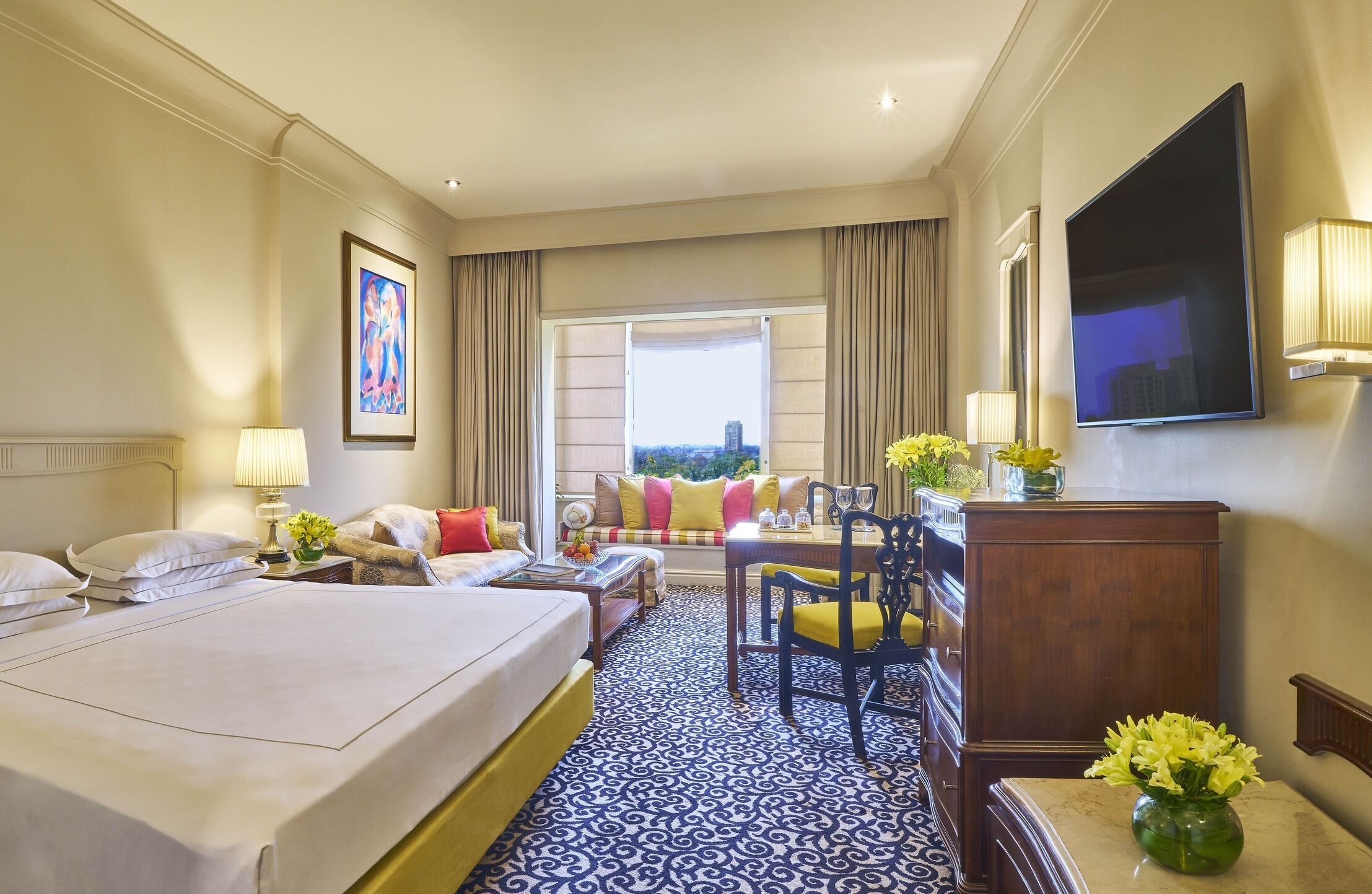 A hotel room with two beds, a sofa, a dining table with chairs, a TV, and a window with a city view.
