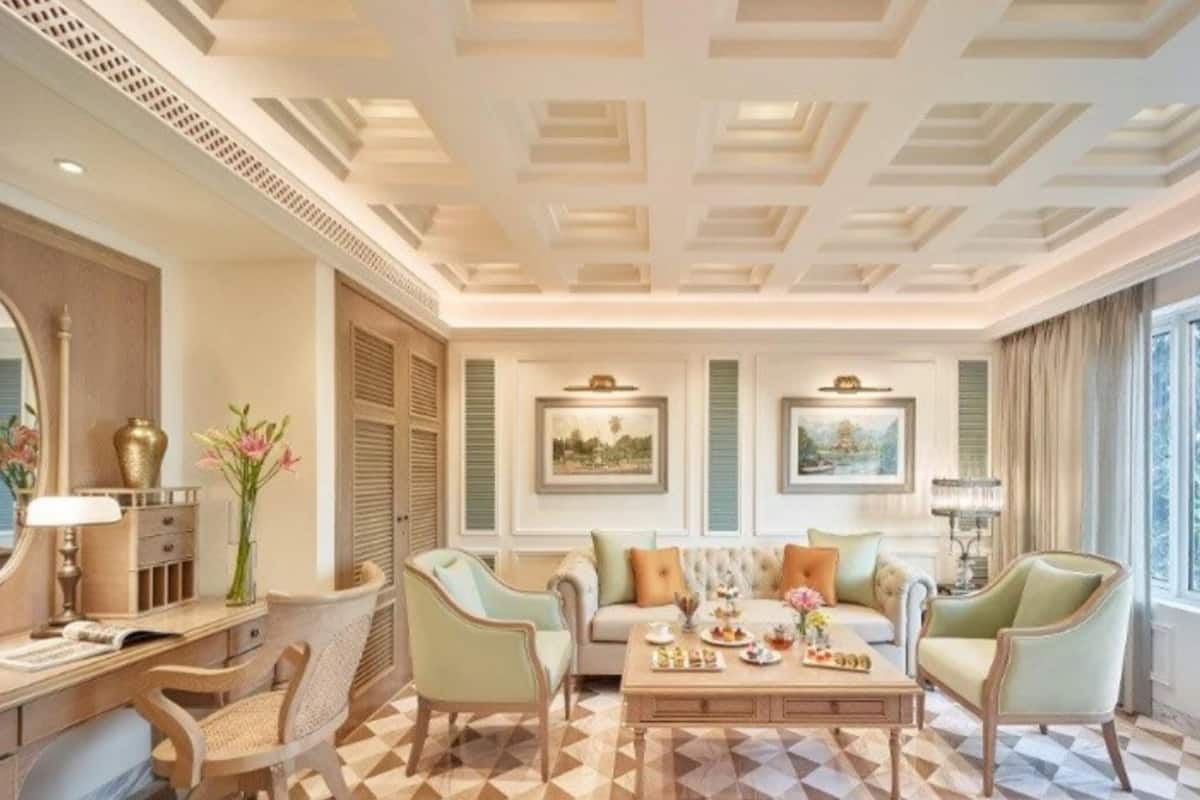 A spacious living room with a checkered floor, a sofa set, a coffee table, and two armchairs. There are two framed artworks on the wall, a large mirror, and a window with curtains.