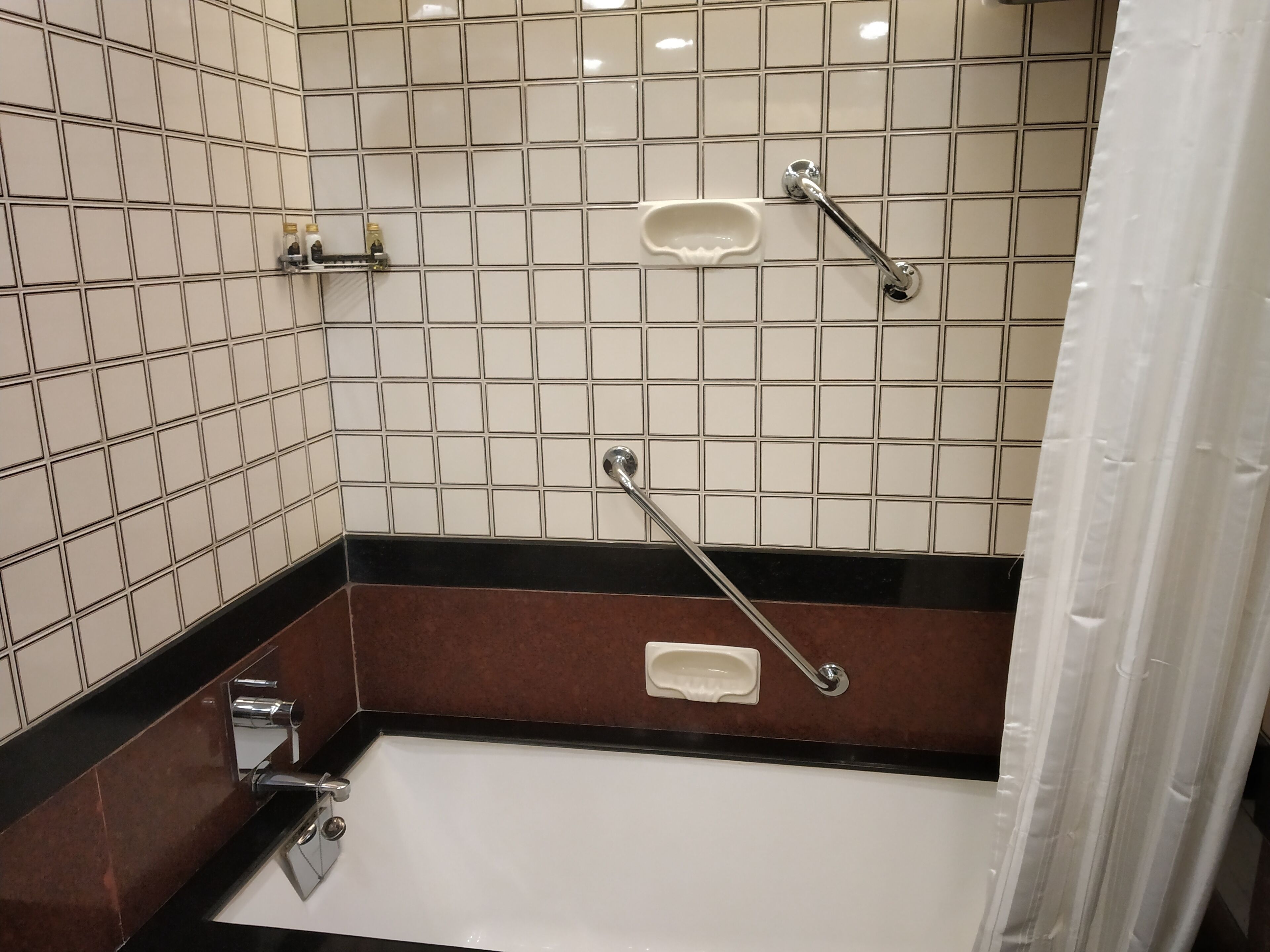A bathroom with white tiled walls, a black and brown lower section, a white bathtub, and a shower area with a grab bar.
