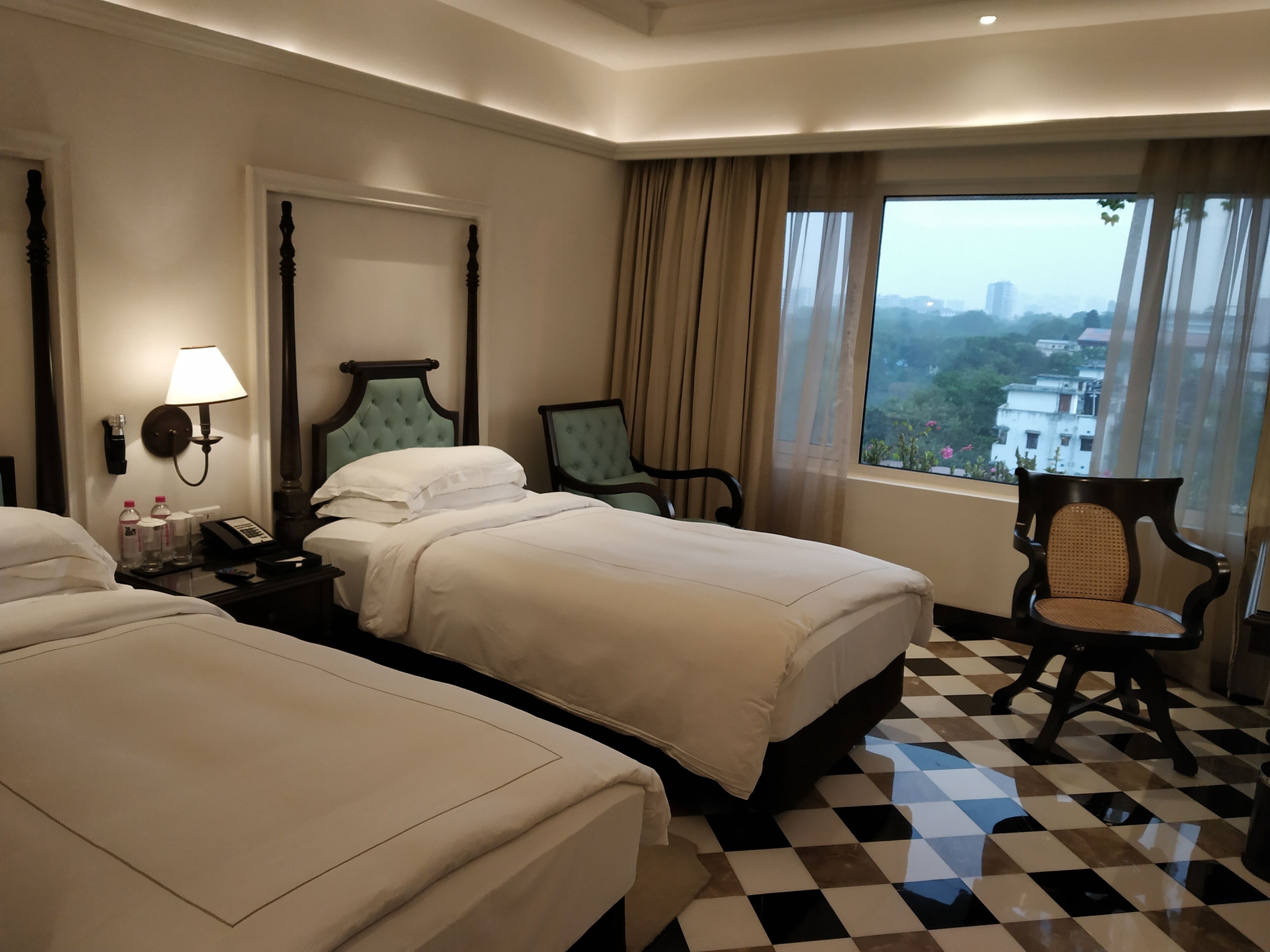 A hotel room with two beds, a nightstand with a lamp, a chair, and a large window with a city view.