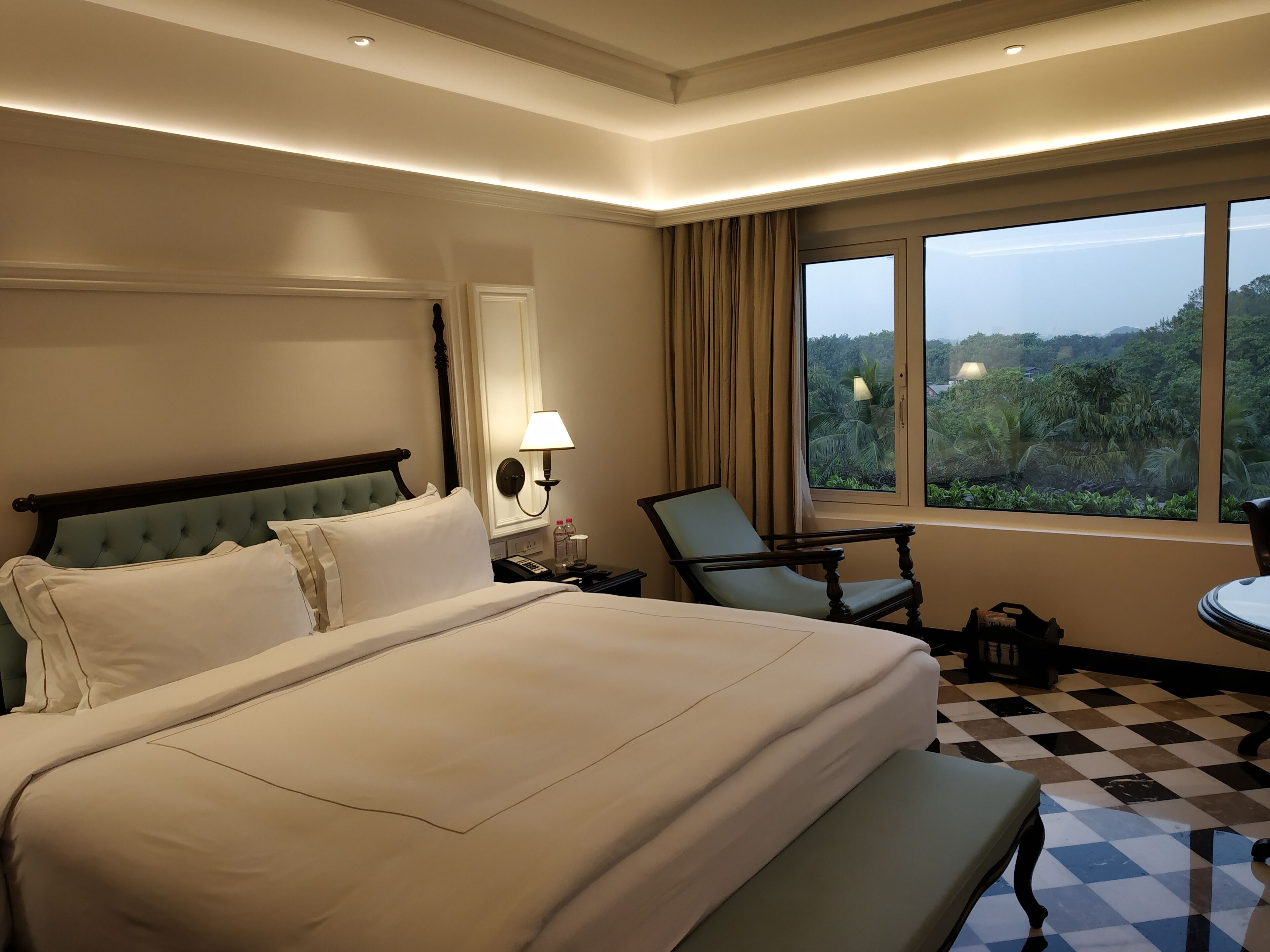 A hotel room with a large bed, a chair, a table, and a window with a view of greenery.