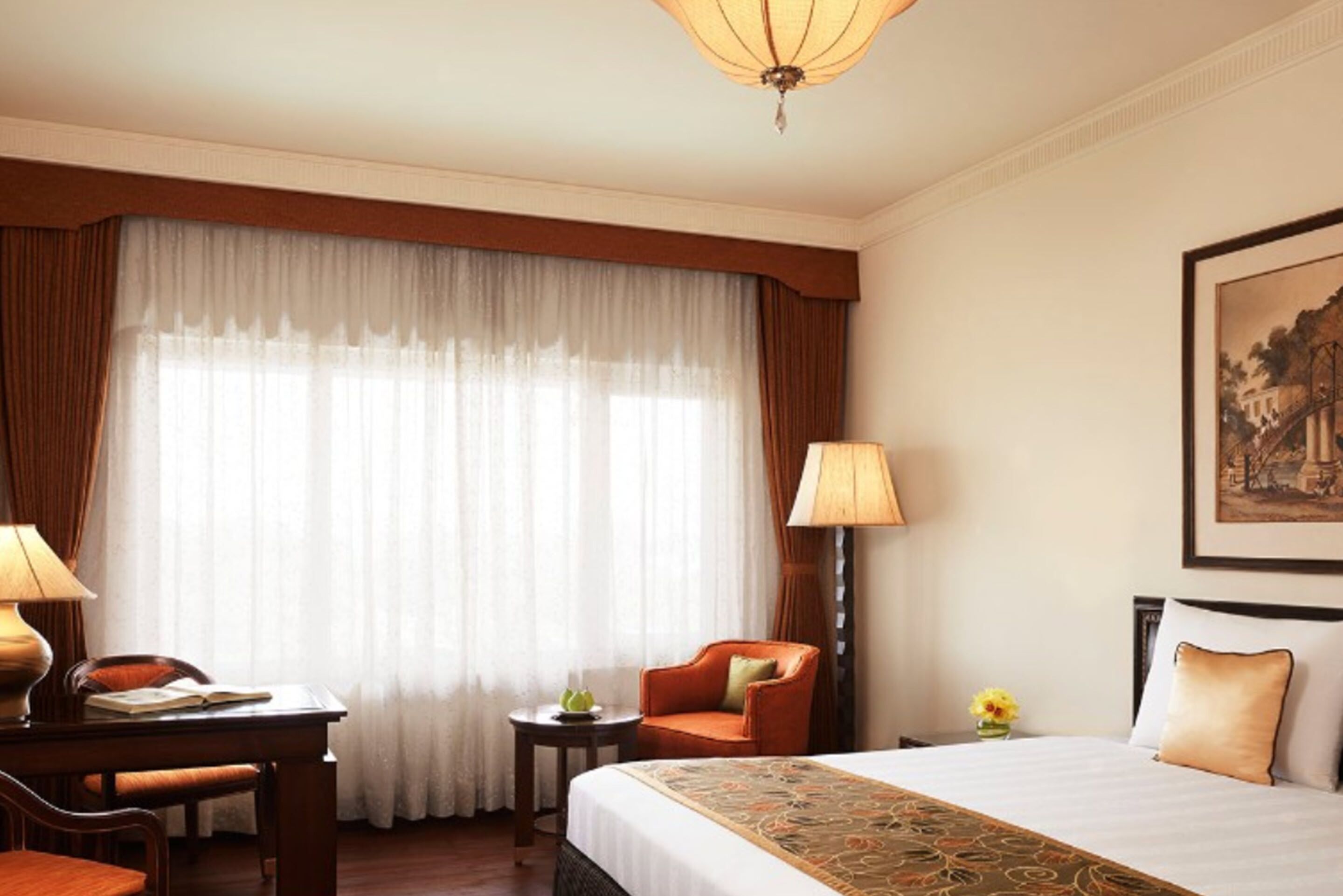 Una habitación de hotel con una cama grande, un escritorio con silla, una mesita de noche con lámpara y un ventanal con cortinas.