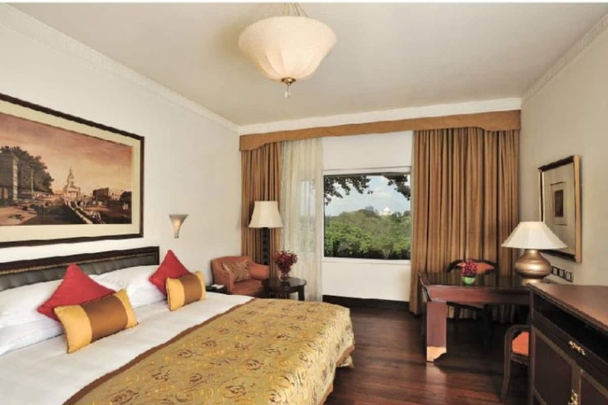 A hotel room with a large bed, a desk, a chair, a lamp, a painting, and a window with curtains.
