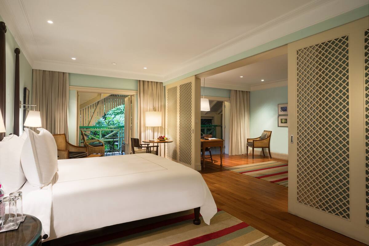 A hotel room with a large bed, a desk, and a chair. There is a balcony with a view of greenery.