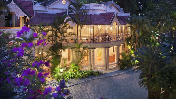 A large, multi-story building with a balcony and a covered patio area, surrounded by lush greenery and palm trees. - Taj West End (Bengaluru)