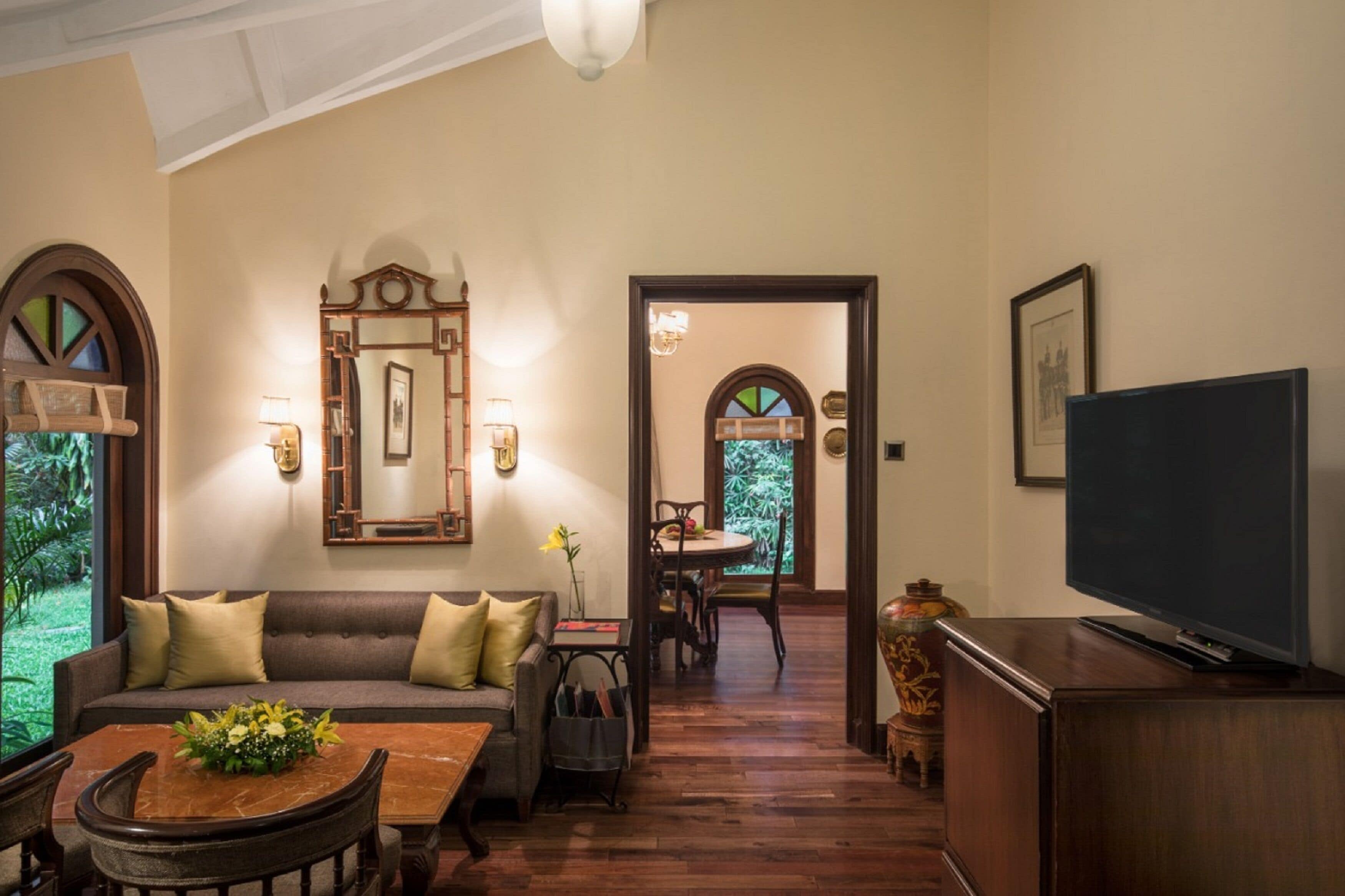 A cozy living room with a sofa, coffee table, and TV. There is a dining area with a table and chairs. The room has wooden floors and a large mirror on the wall.
