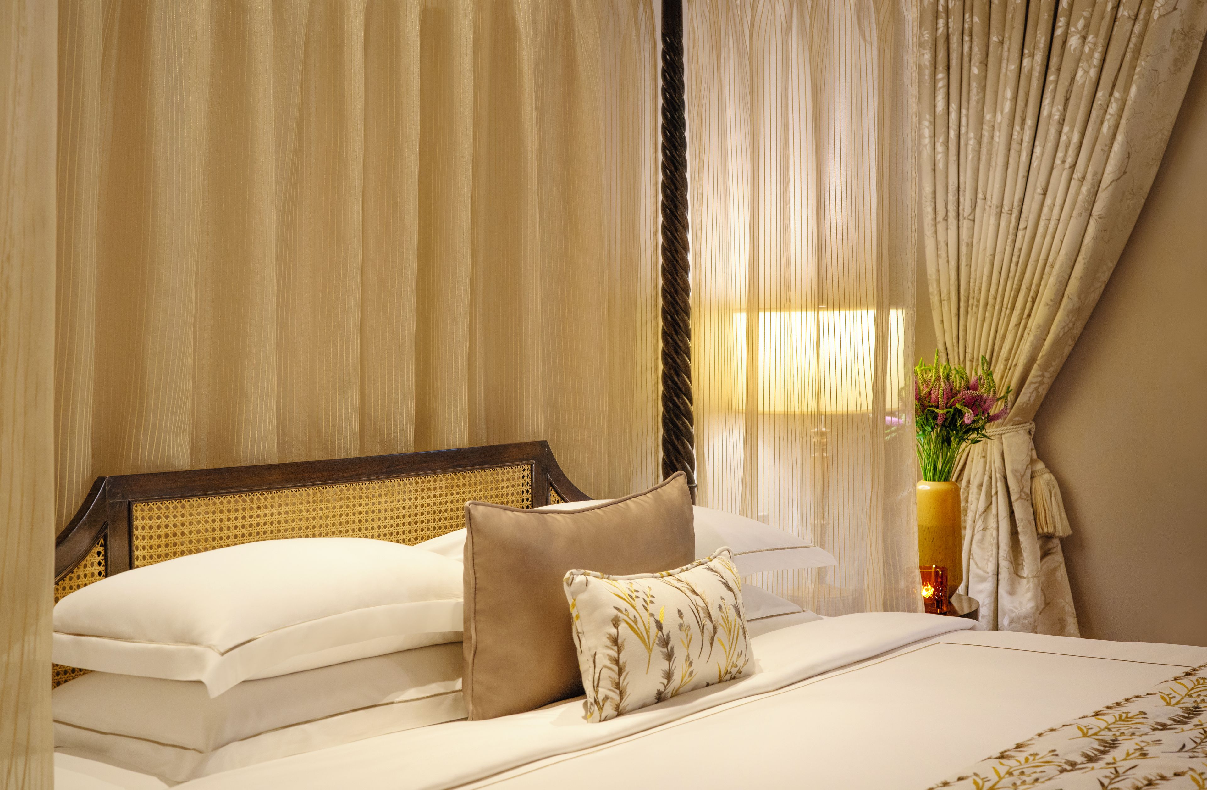 Una habitación de hotel con cama, cortinas, un florero con flores y una mesita de noche.