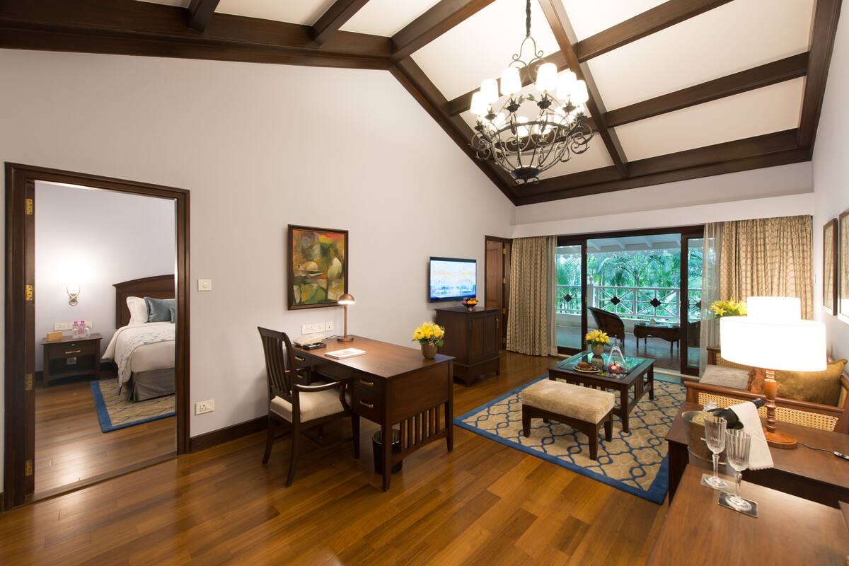 A hotel room with a wooden floor, a desk with a chair, a dining table with chairs, a sofa, a TV, and a large window with curtains.