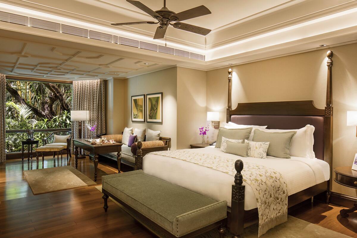 A spacious bedroom with a large bed, a sitting area with a chair and table, and a balcony with a view of greenery.