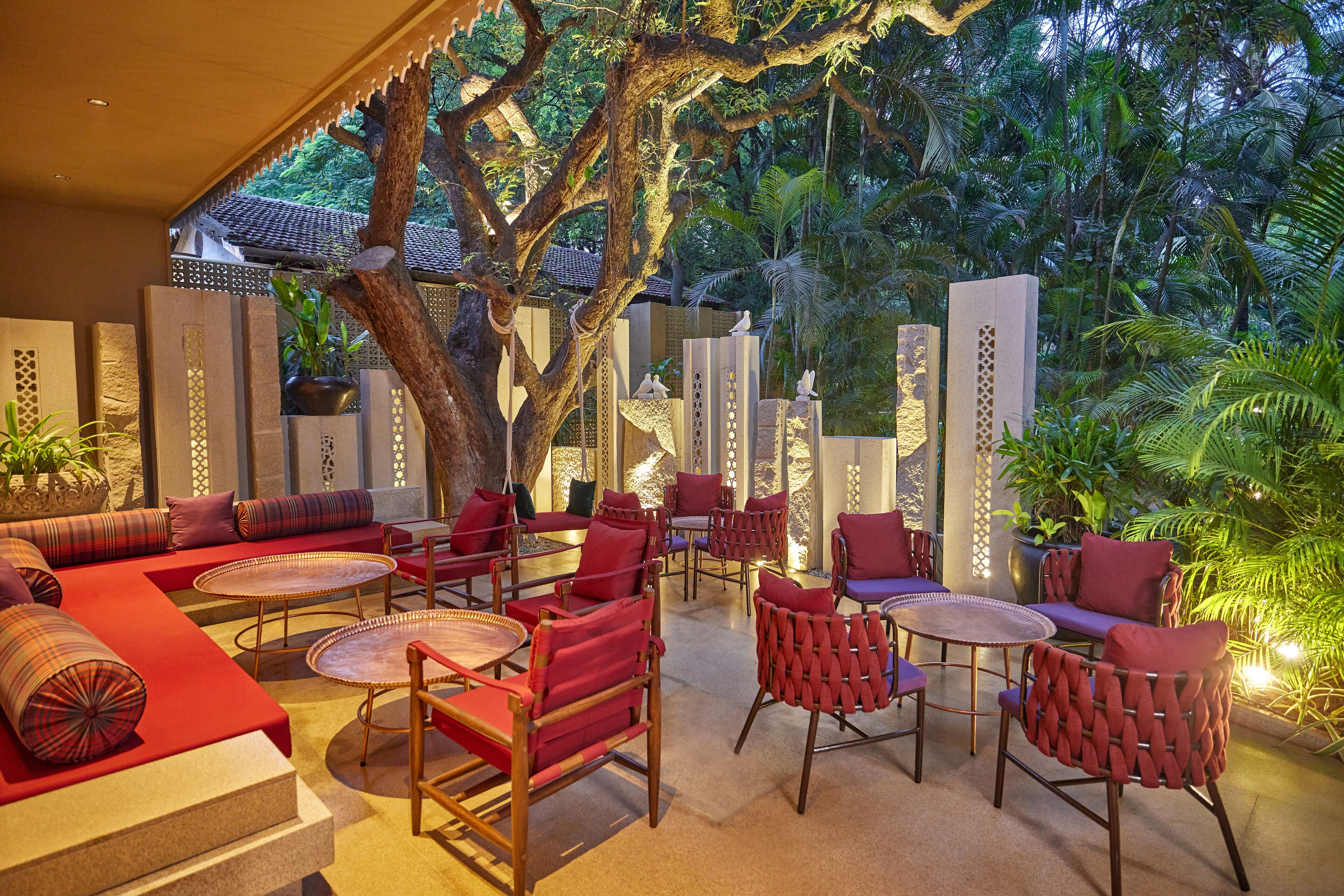 Un espacio al aire libre con mobiliario tapizado en rojo, mesas redondas y un gran árbol que da sombra.