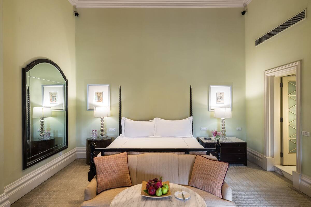 A hotel room with a large bed, two bedside tables with lamps, a round coffee table with fruit, and a sofa with cushions.