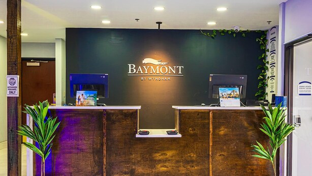 A reception desk with a dark wooden finish, a logo above it, and two potted plants on either side. - Baymont by Wyndham Port Wentworth (Port Wentworth)