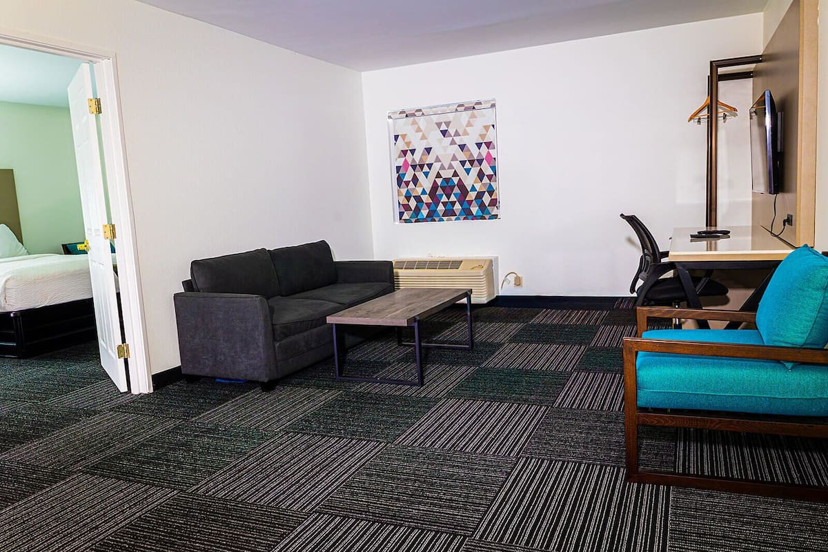 A hotel room with a bed, a gray sofa, a wooden coffee table, a teal armchair, a desk with a computer, and a wall-mounted TV.