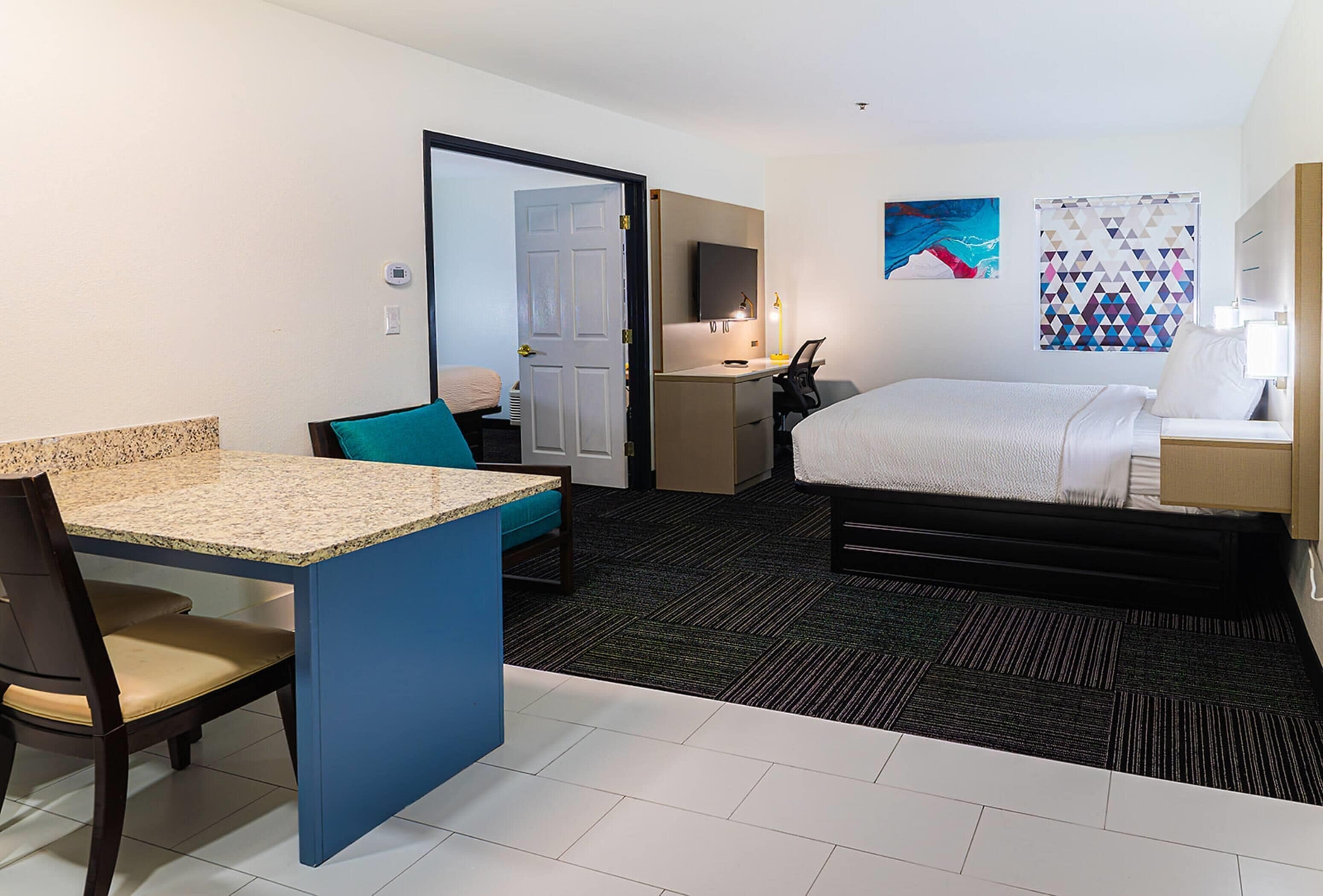 Habitación de hotel con cama, escritorio, silla, kitchenette pequeña y baño.