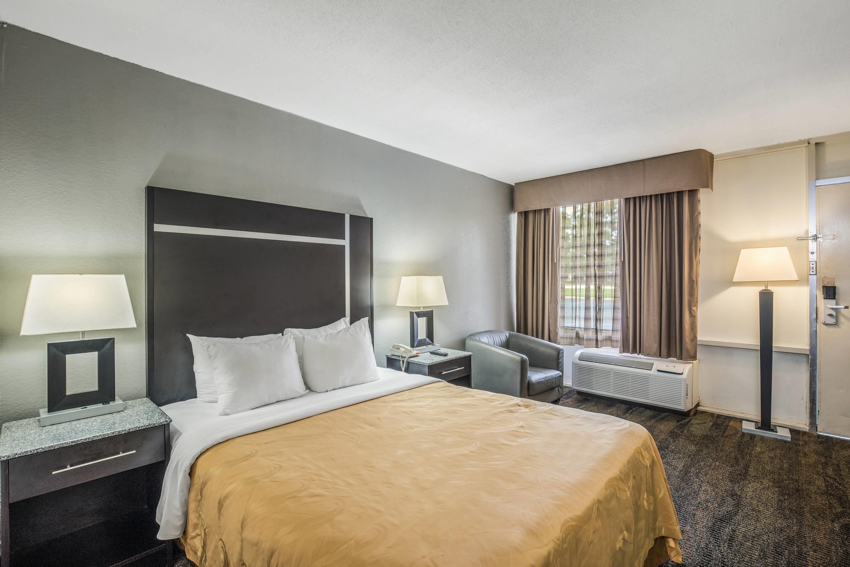 Une chambre d’hôtel comprenant un lit, deux tables de chevet, une chaise, une fenêtre avec des rideaux et un radiateur.