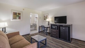 A hotel room with a sofa, a desk with a chair, a television, a refrigerator, and a bathroom visible through an open door. - Quality Inn & Suites Conference Center (Thomasville)