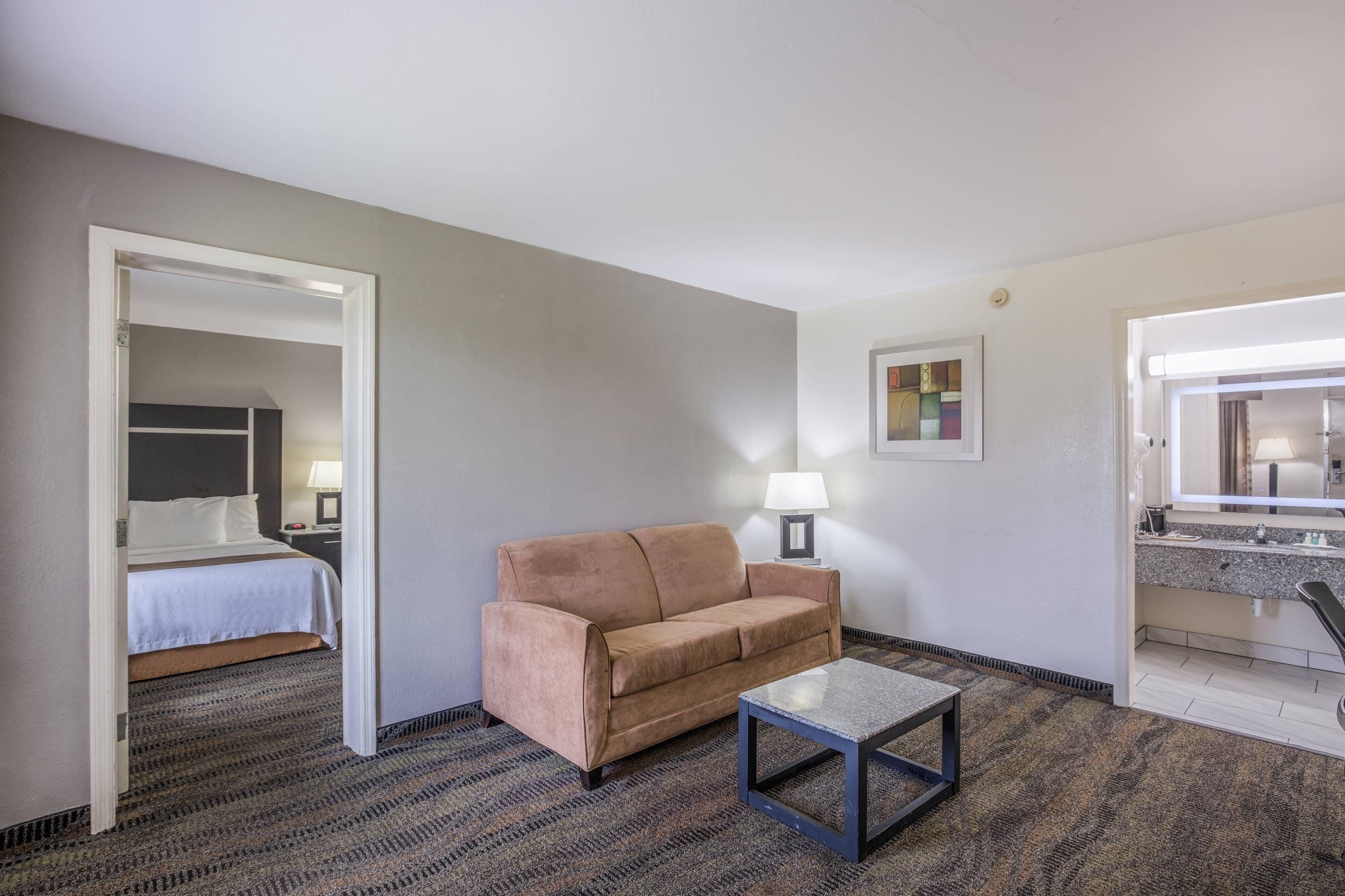 Habitación de hotel con cama, sofá, mesa de centro, lámpara, un cuadro y baño visible a través de una puerta abierta.