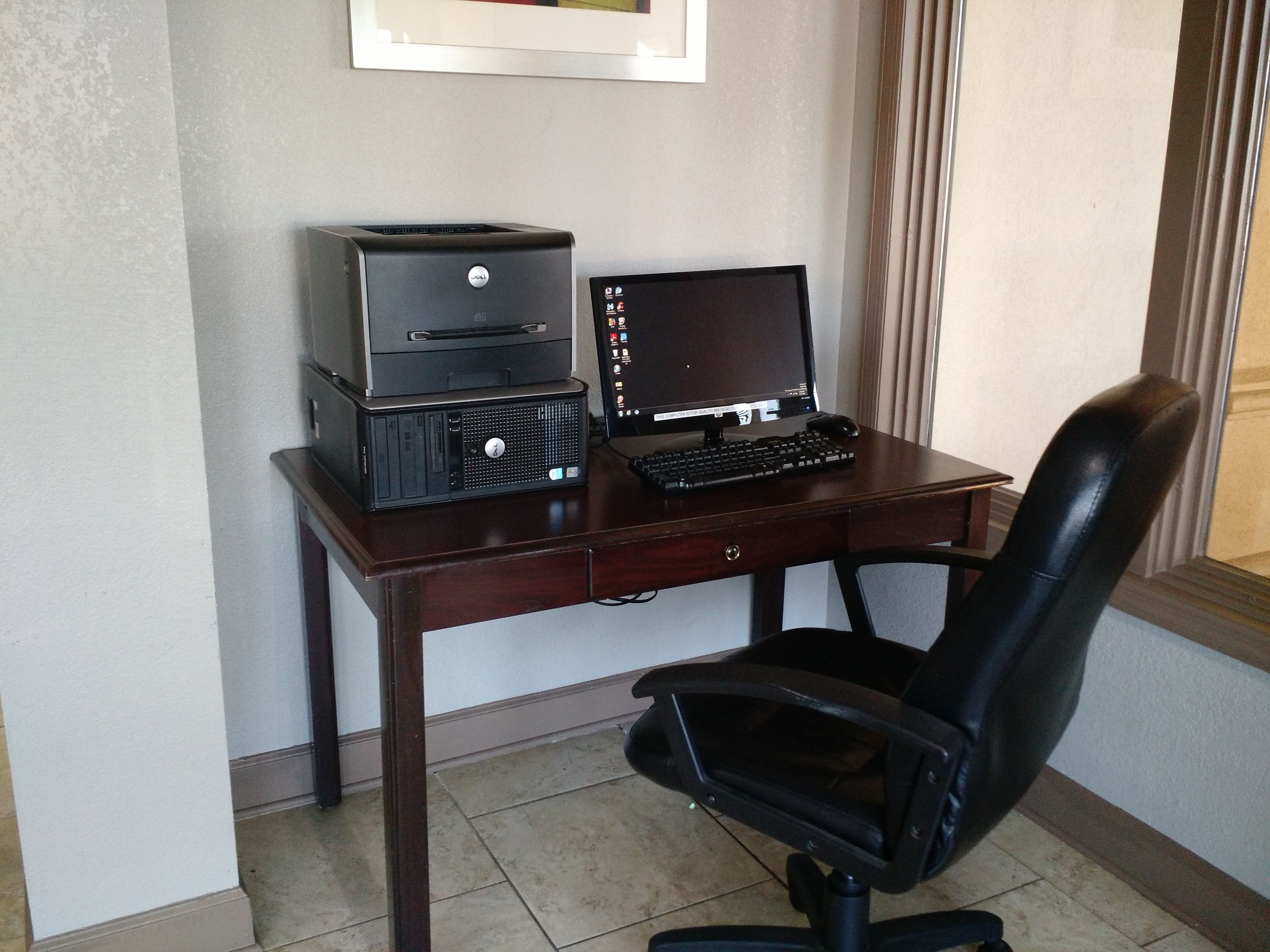 Een houten bureau met een desktopcomputer, een printer en een zwarte kantoorstoel.