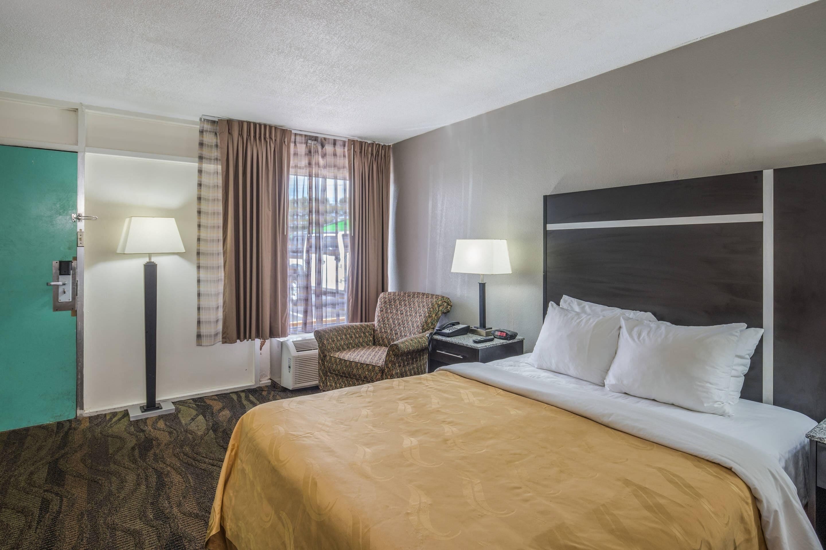 Quarto de hotel com cama, duas luminárias de cabeceira, uma cadeira, escrivaninha com telefone e janela com cortinas.