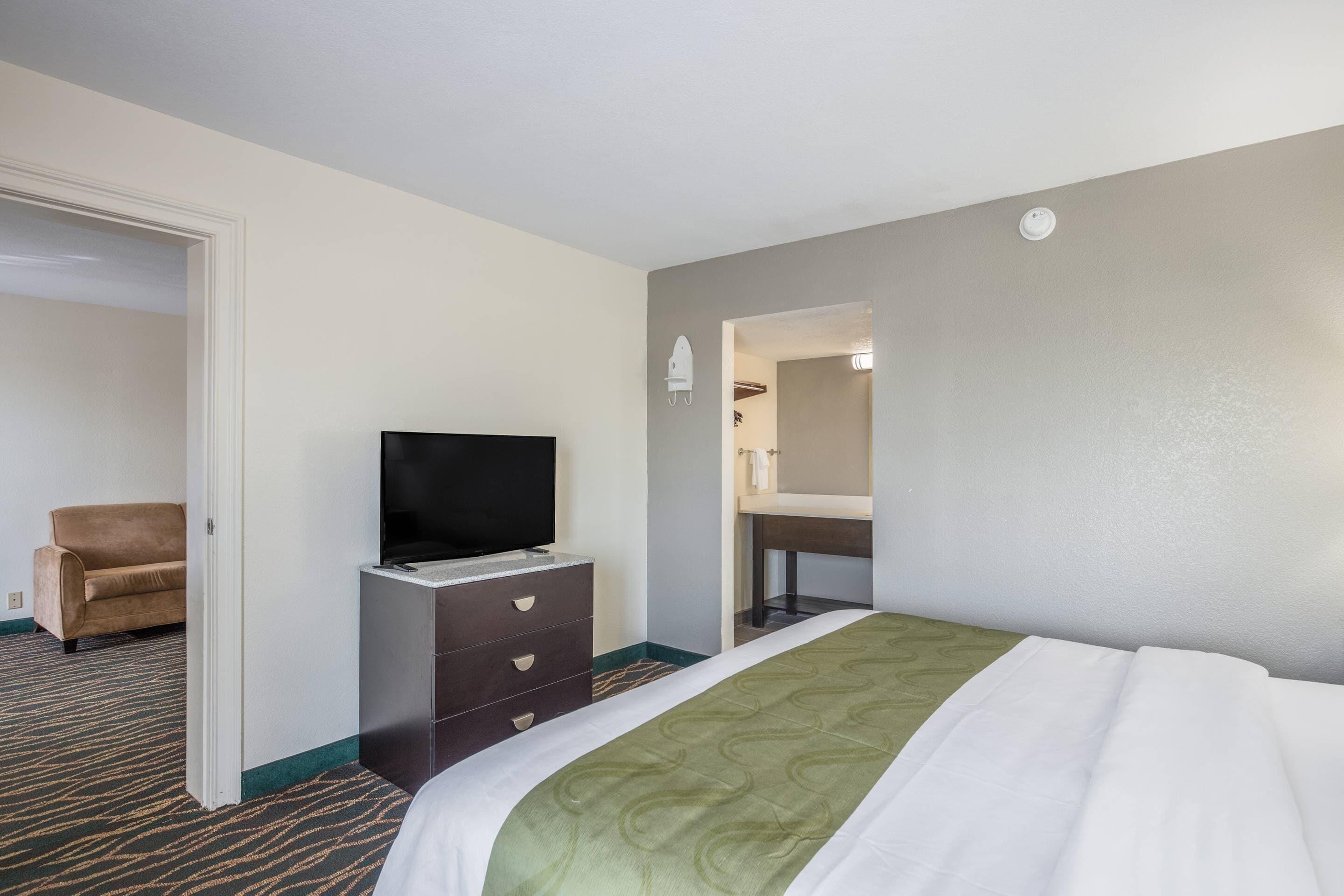 A hotel room with a bed, a TV on a dresser, a chair, and a bathroom visible through an open door.