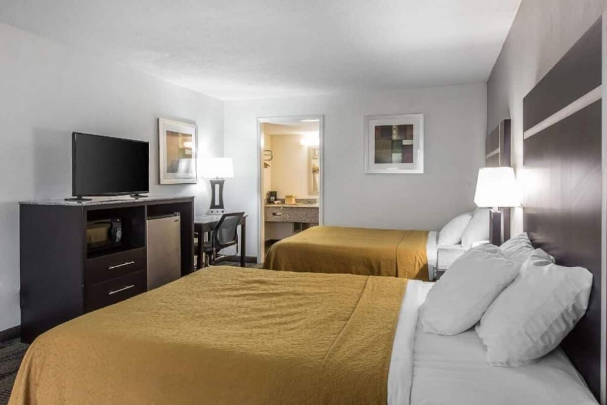 Une chambre d’hôtel avec deux lits, un bureau, une télévision et une salle de bain visible à travers une porte ouverte.