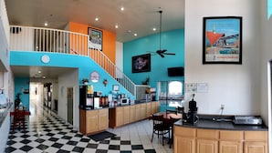 A hotel lobby with a checkered floor, a staircase, a kitchen area with a coffee machine, and a dining area with chairs.