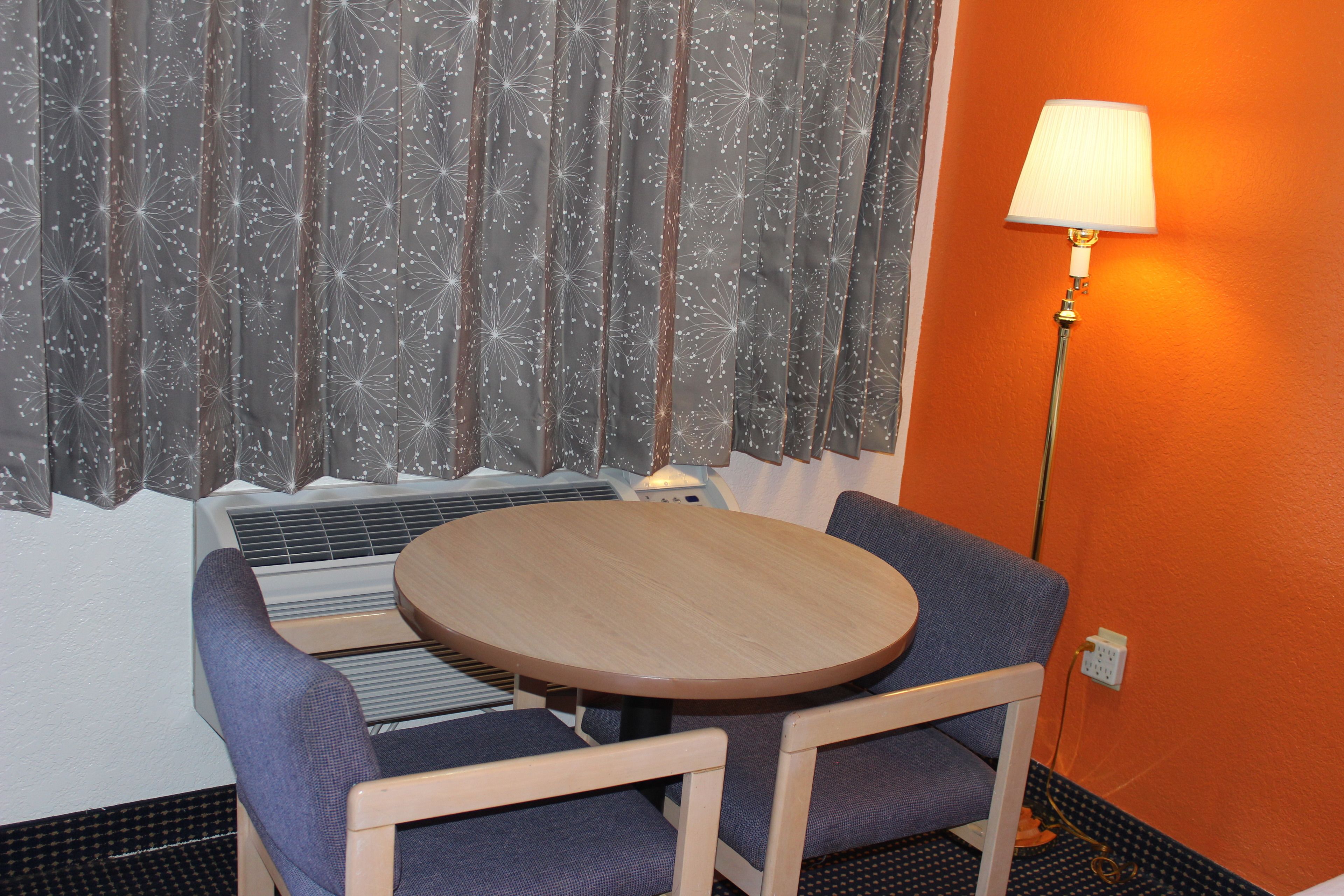 Um quarto com uma mesa redonda de madeira, duas cadeiras azuis, um abajur e uma janela com cortina.