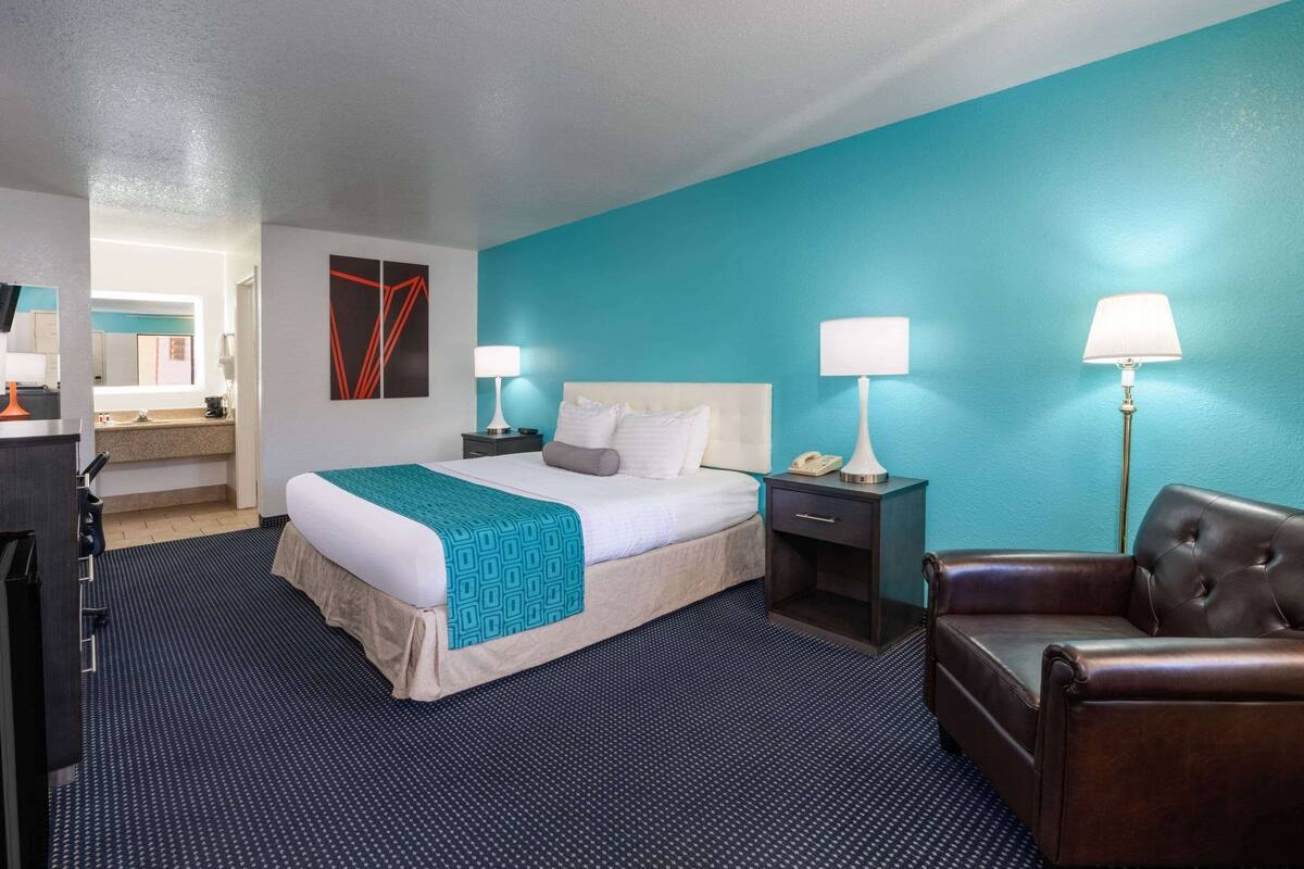 A hotel room with a bed, bedside tables, a chair, and a bathroom visible in the background.