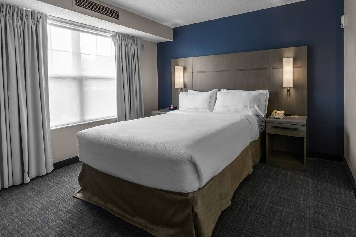 A hotel room with a large bed, a nightstand with a lamp, a telephone, and a window with curtains.