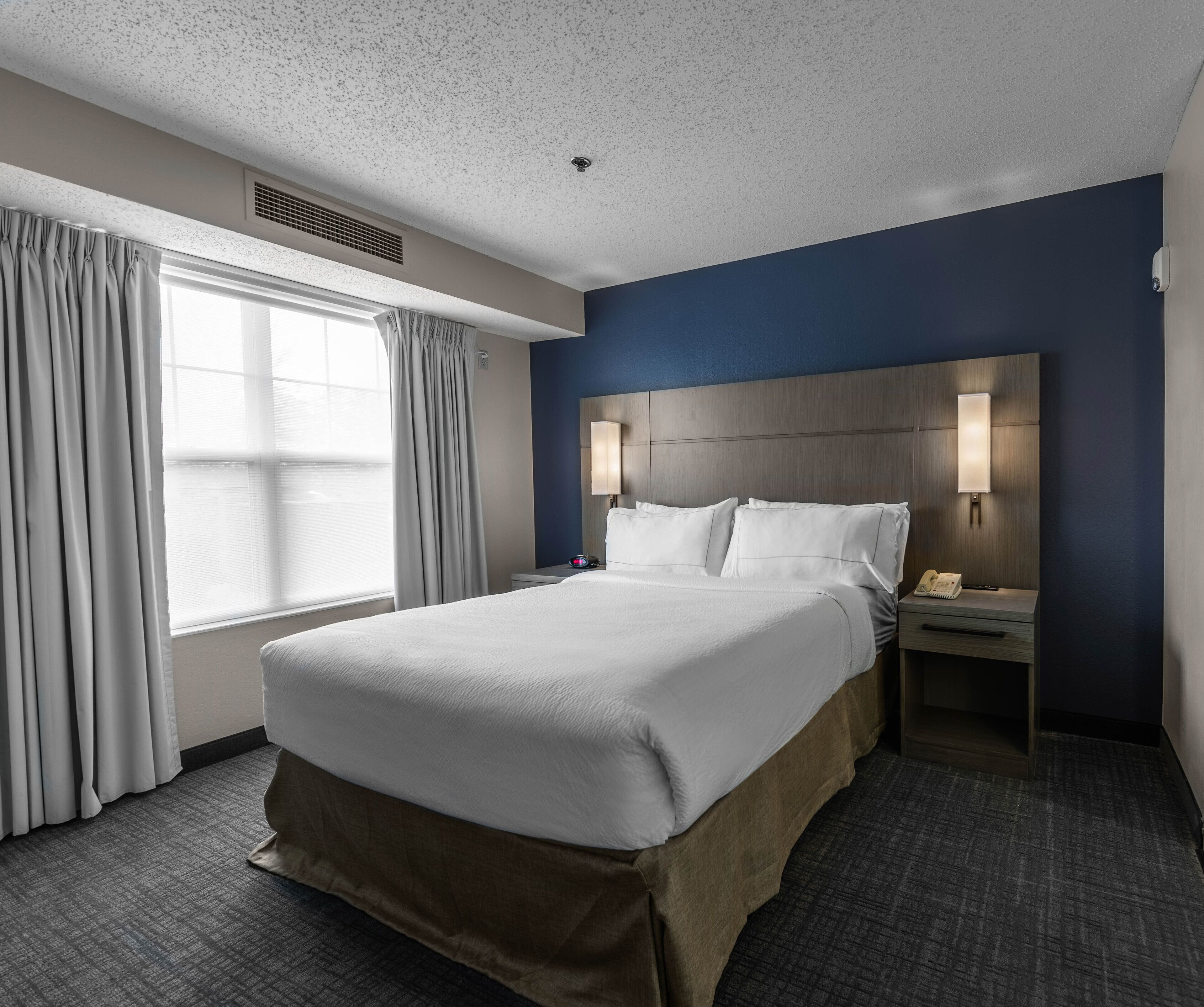 Een hotelkamer met een groot bed, een nachtkastje met een lamp, een telefoon en een raam met gordijnen.